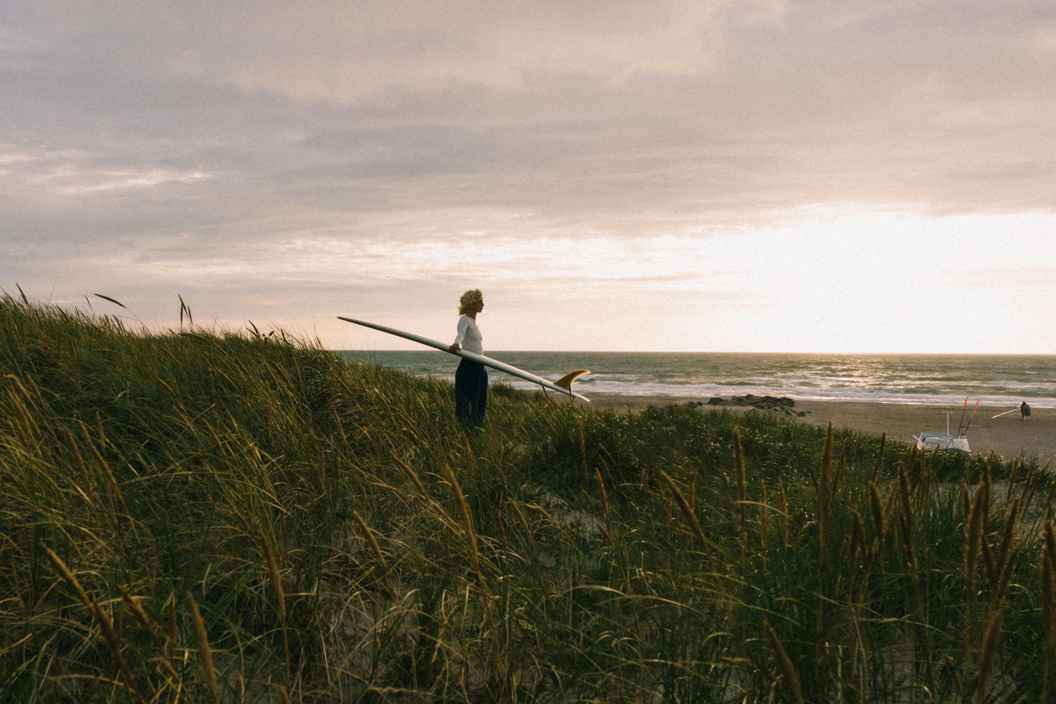 Person holding a surfboard standing on a grassy dune overlooking the ocean at sunset