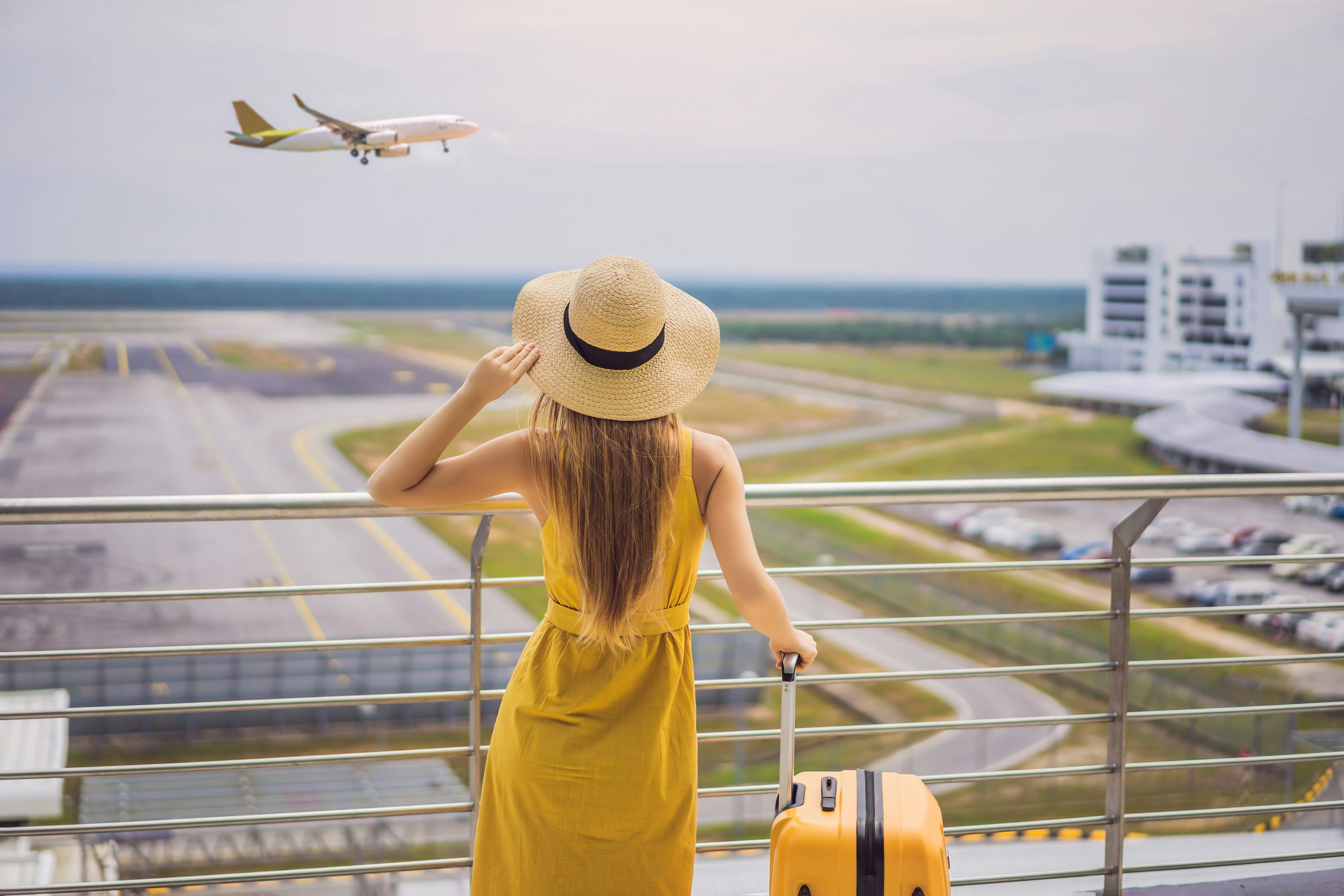Woman in yellow dress with suitcase watching airplane take off at airport
