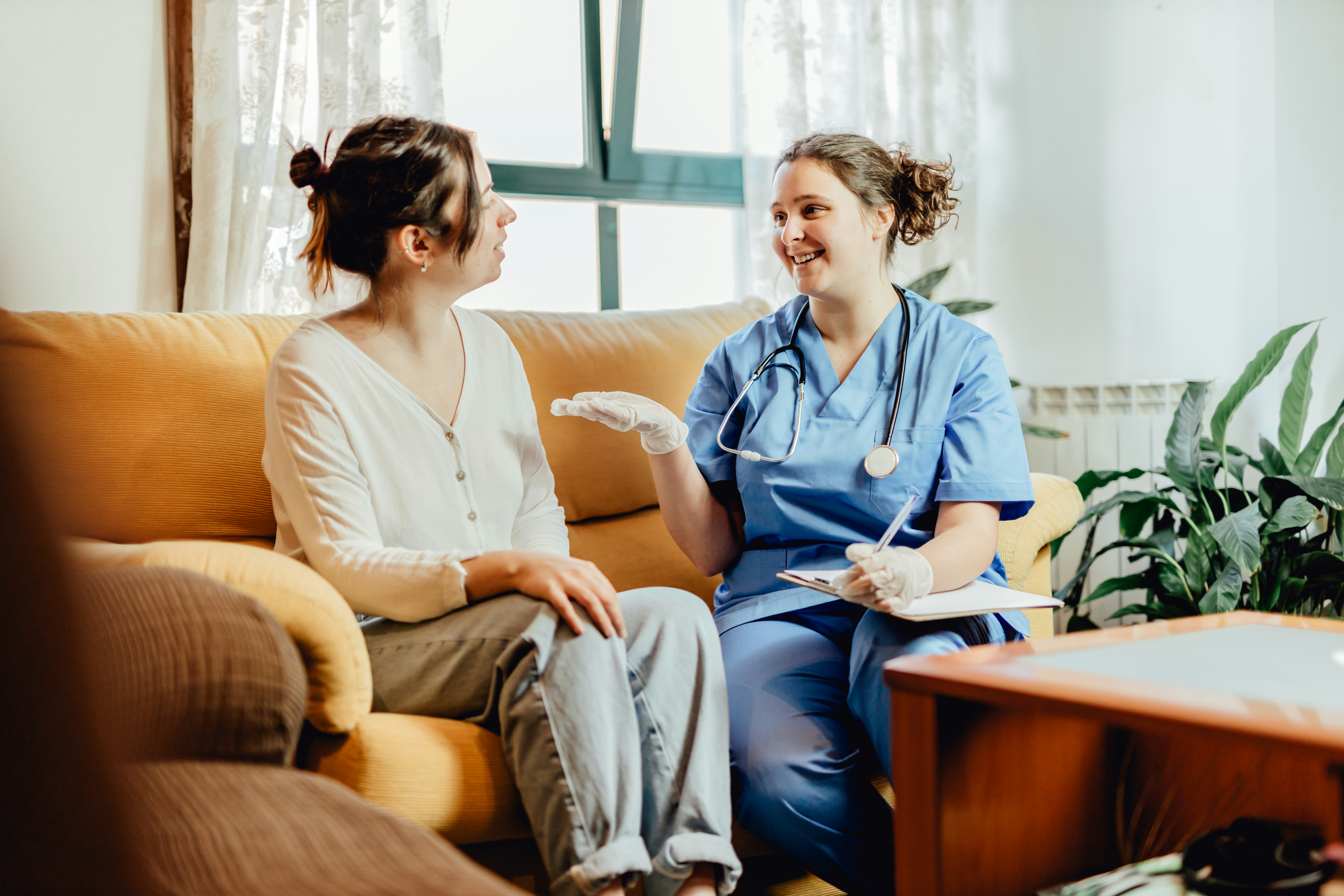 Healthcare professional in scrubs talking to a seated woman in a living room setting.