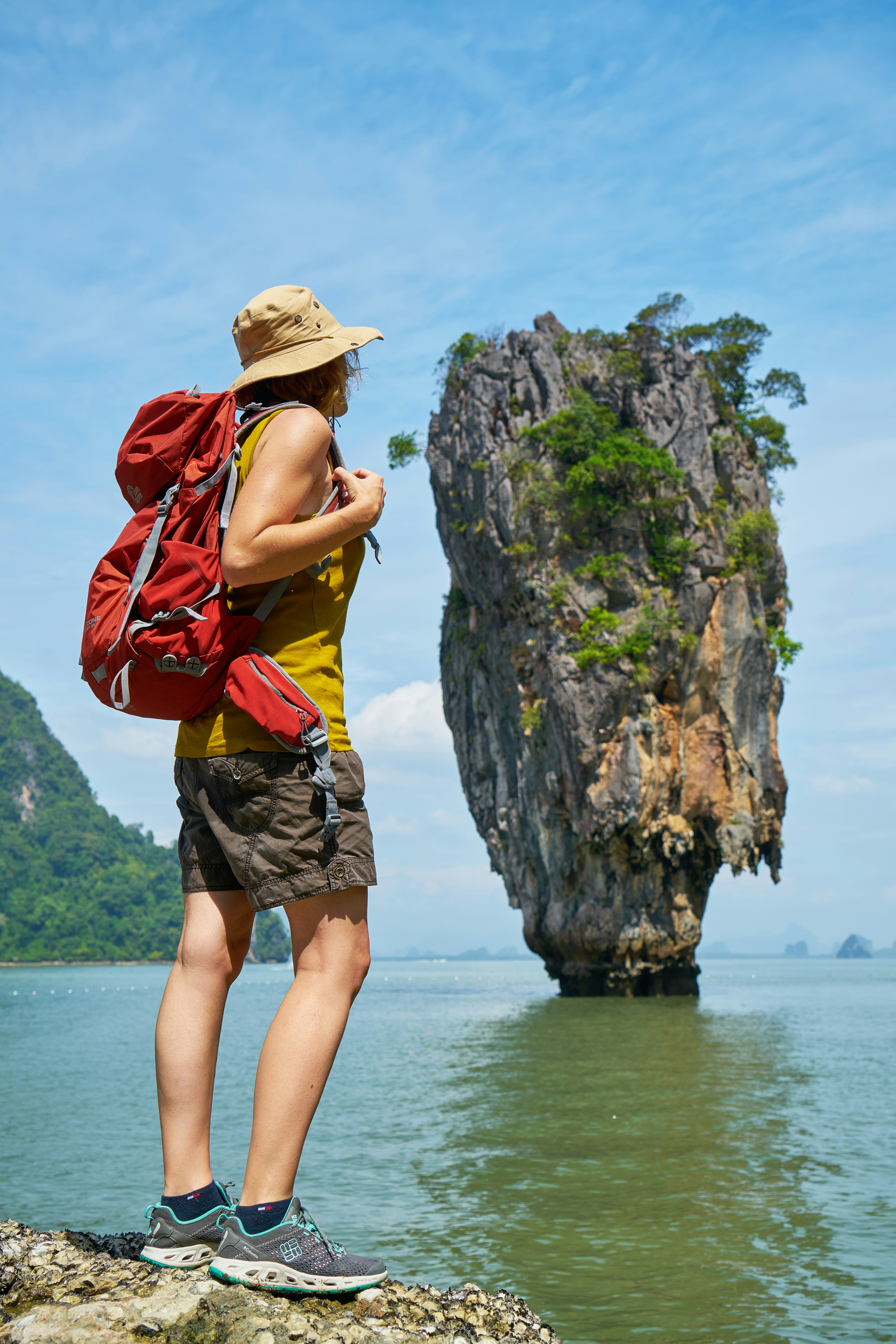Woman with a backpack standing on a rocky shore looking at a tall limestone island in the sea.