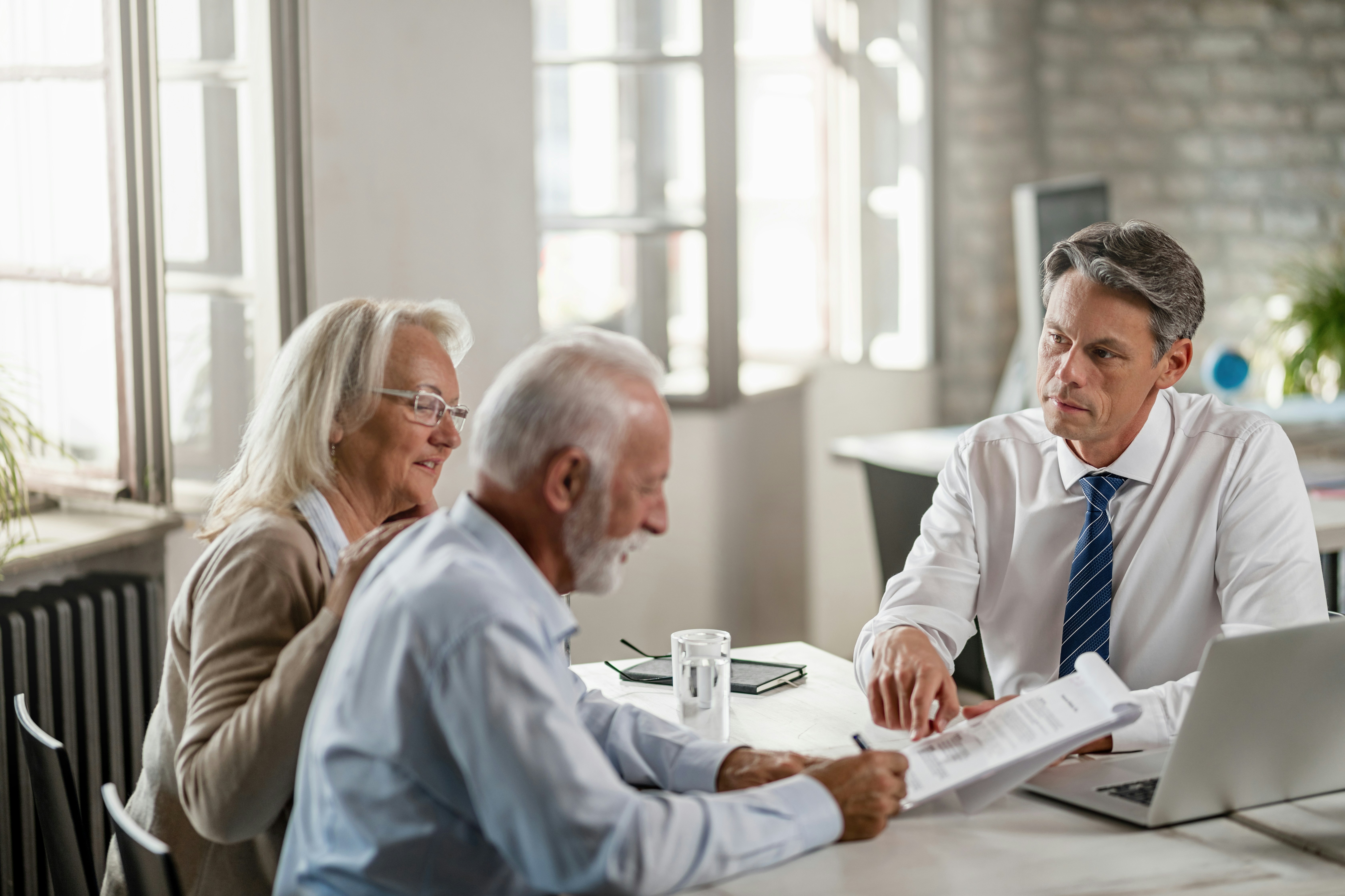 Three coworkers review documents at a table, smiling in a bright office meeting room.