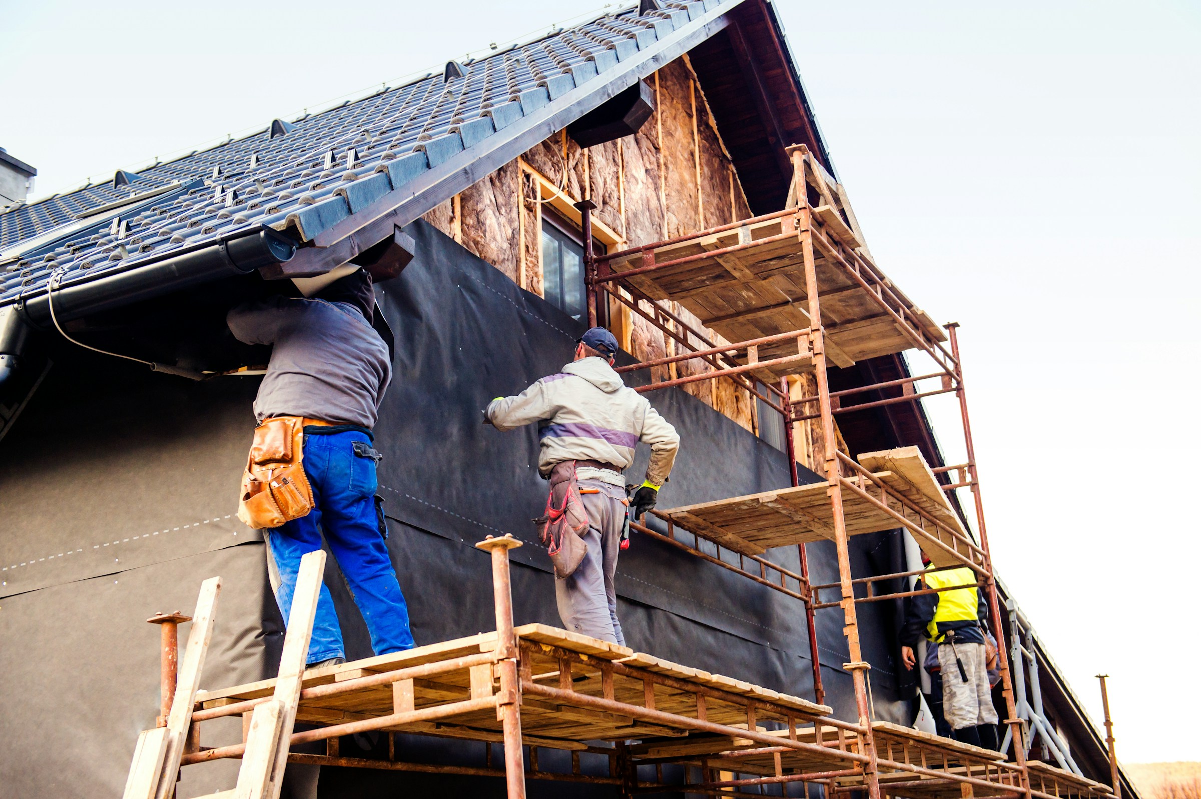 Construction workers installing roofing and siding on a house with scaffolding.