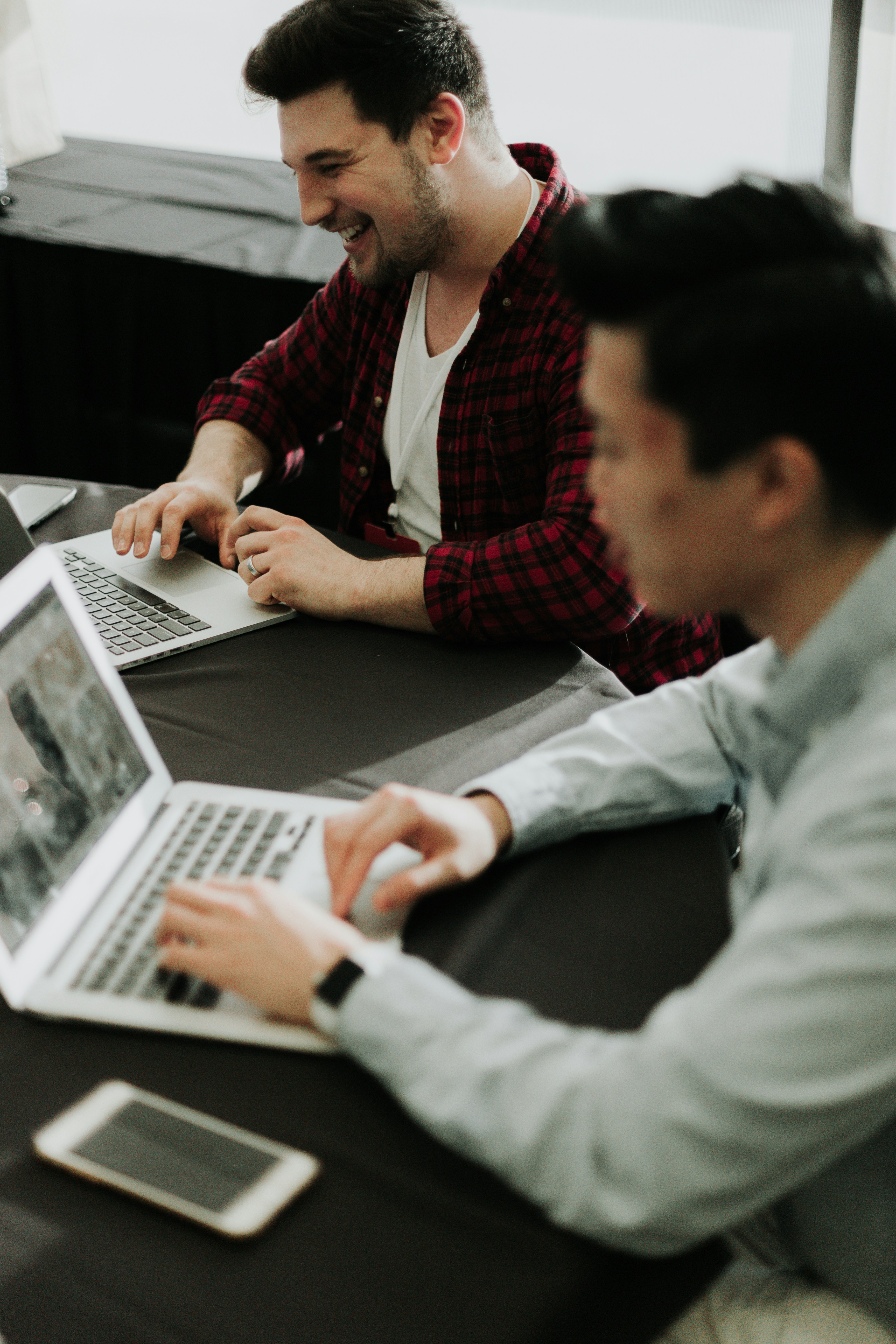 Two men working on laptops at a table with a smartphone nearby