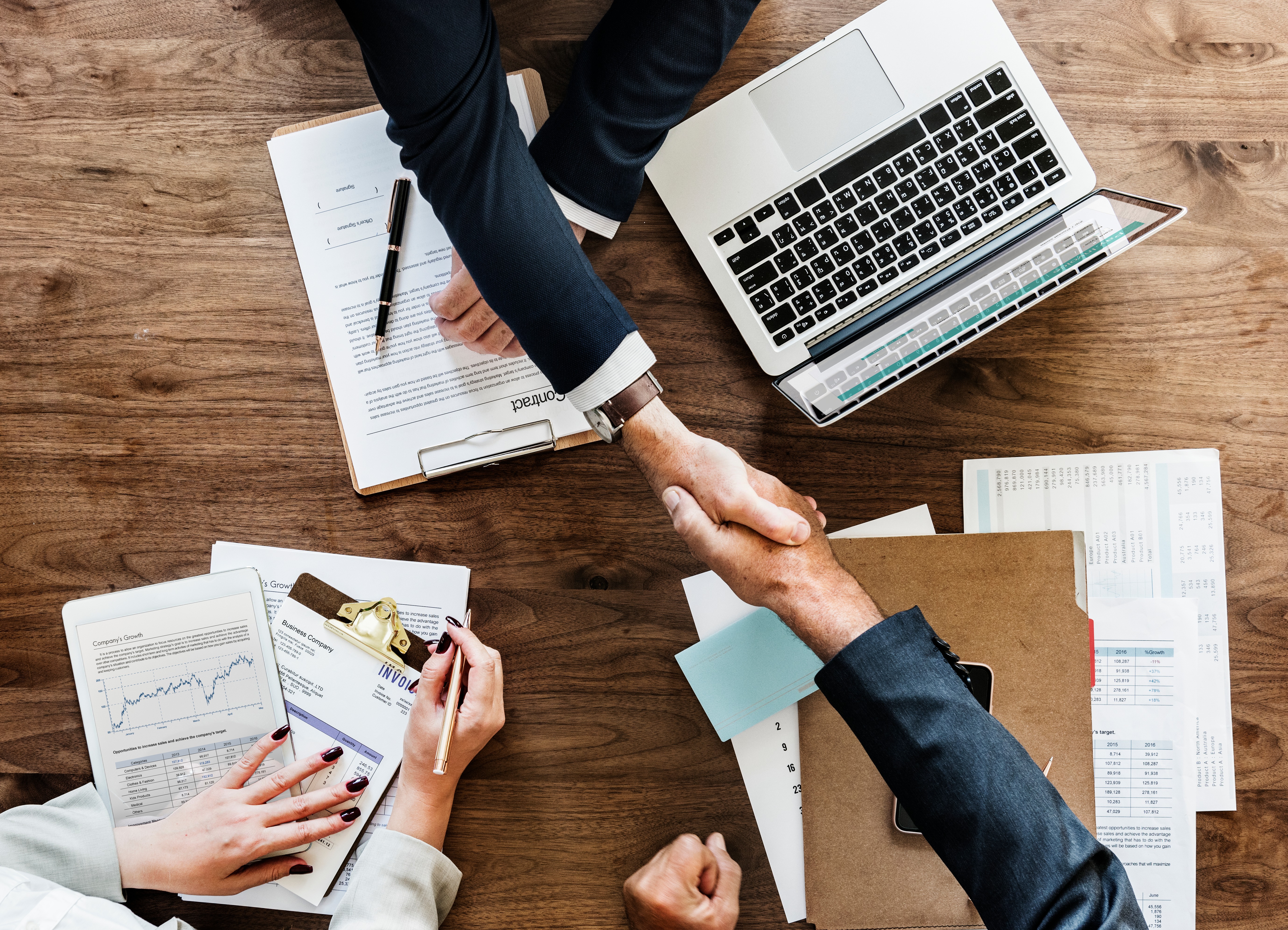 Business professionals shaking hands over a desk with documents and a laptop