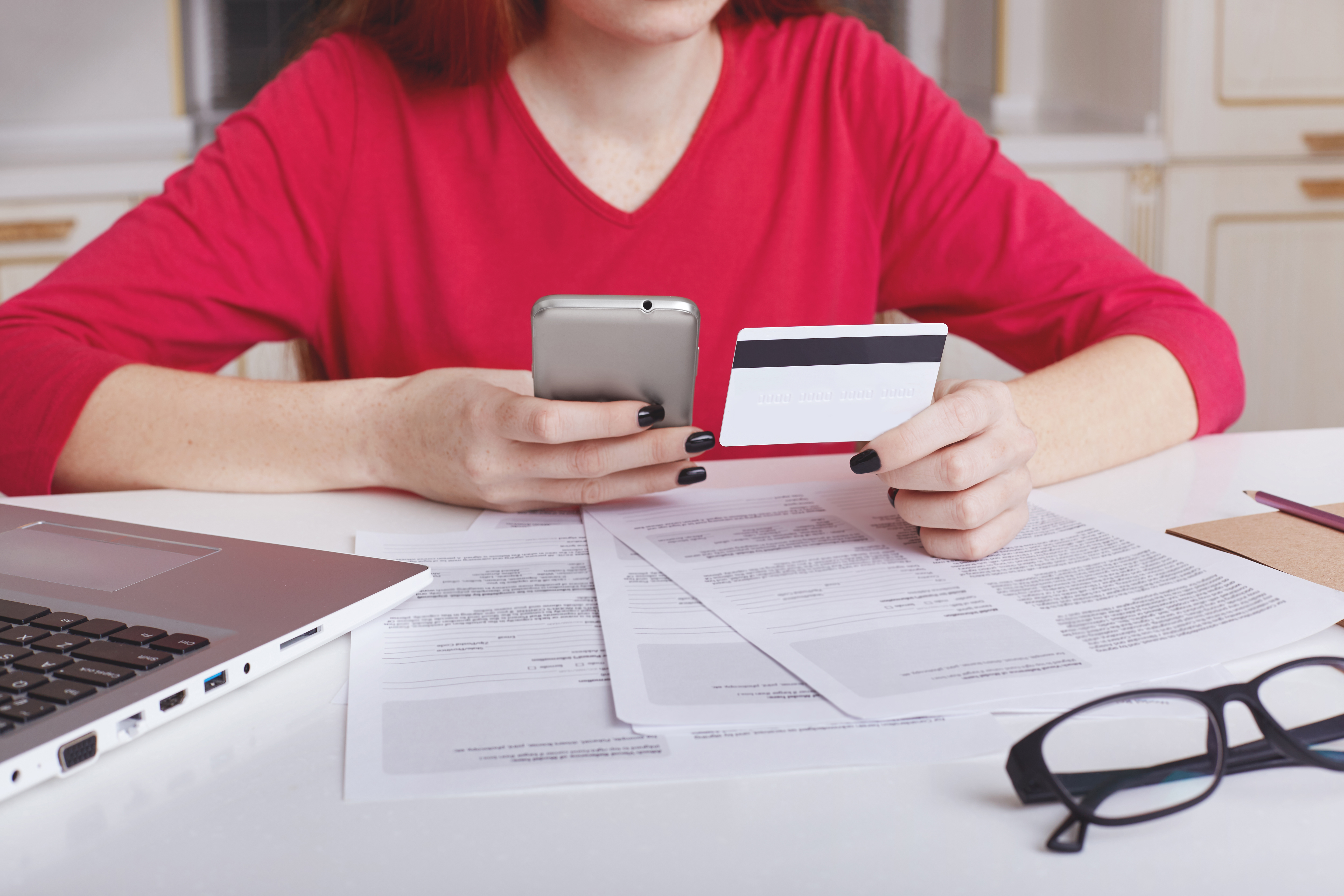 Person in red shirt holding a credit card and smartphone over financial documents.