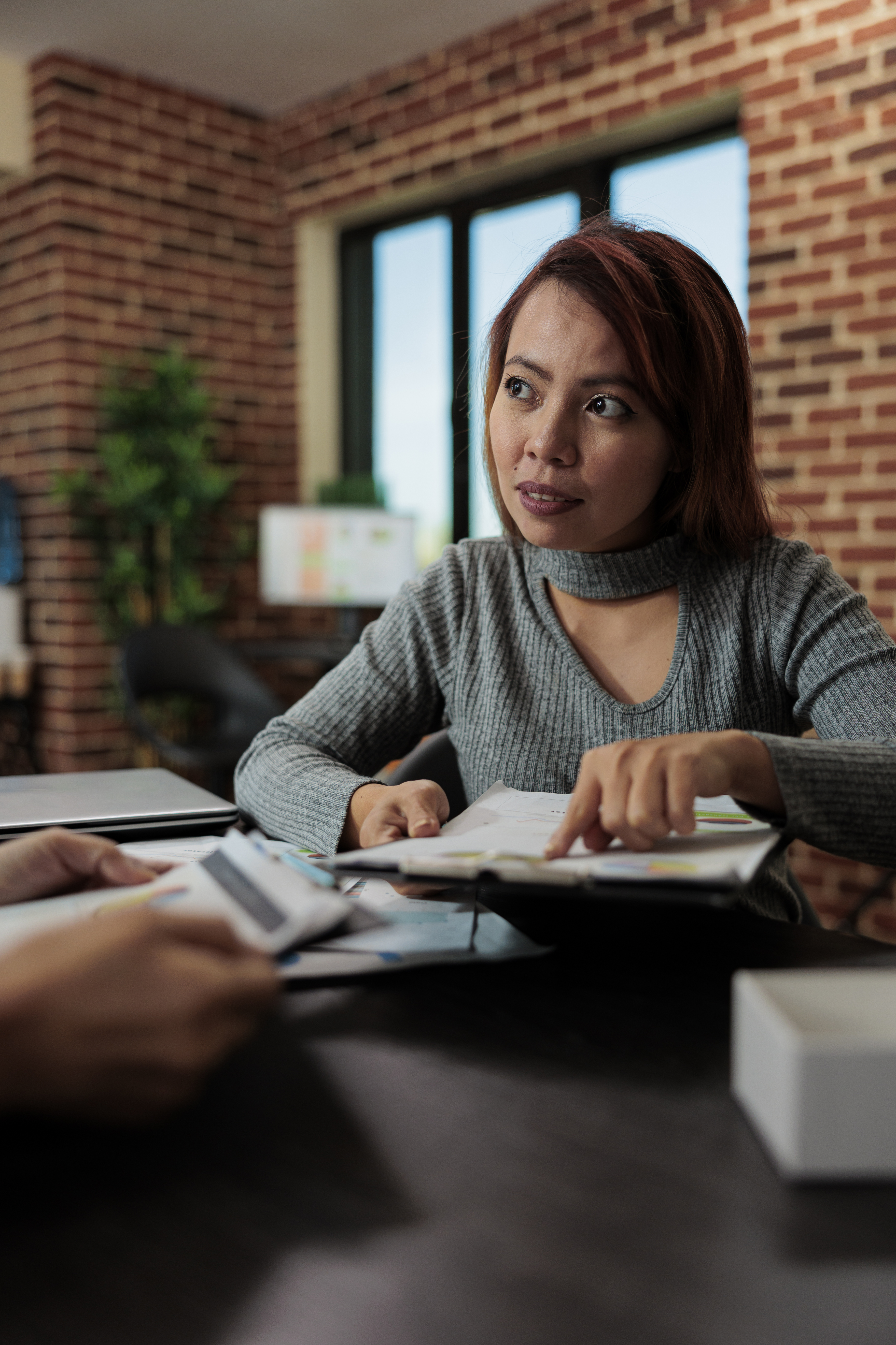 Woman in a gray sweater discussing documents at a desk in an office.