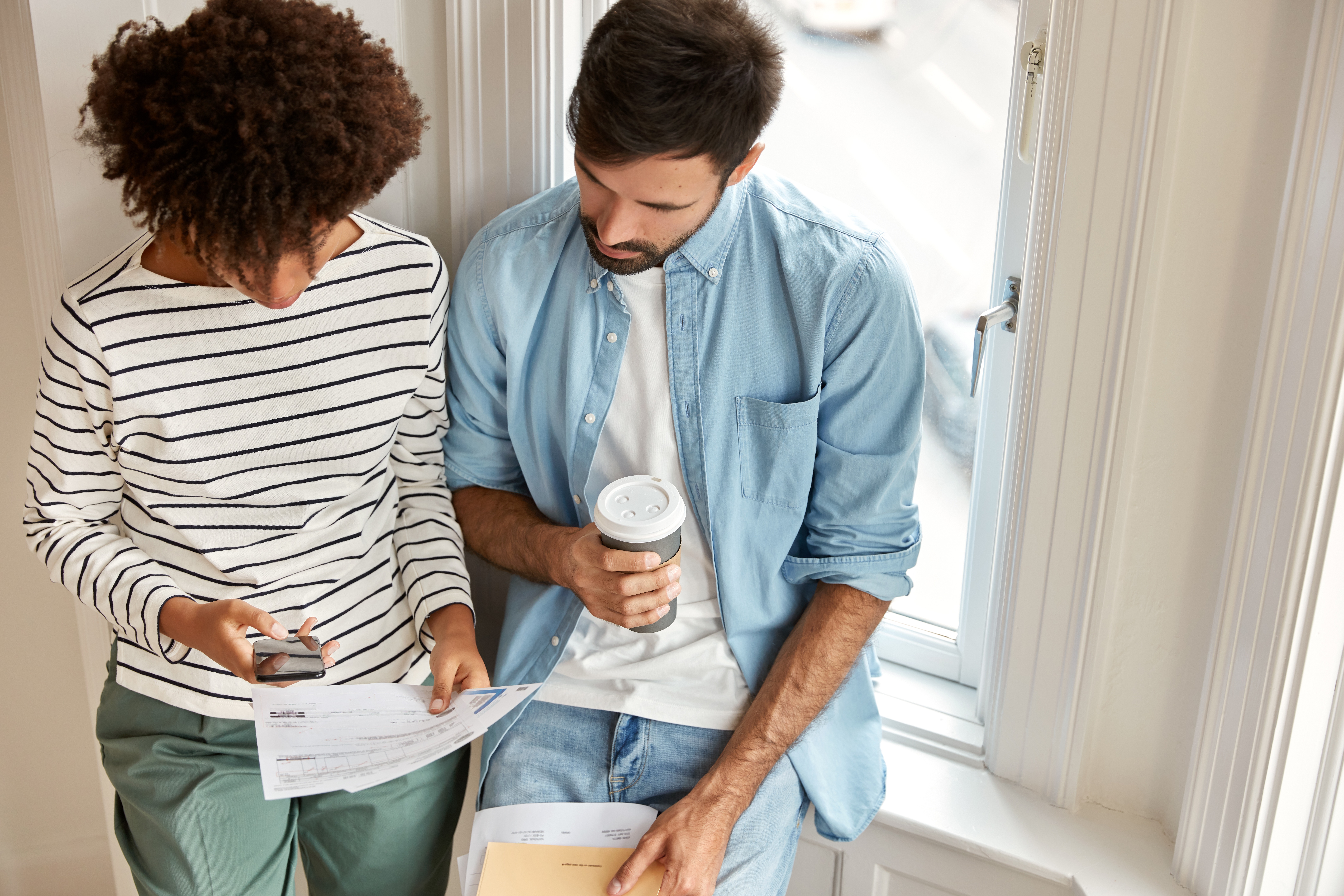 Two people reviewing documents by a window, one holding a coffee cup.