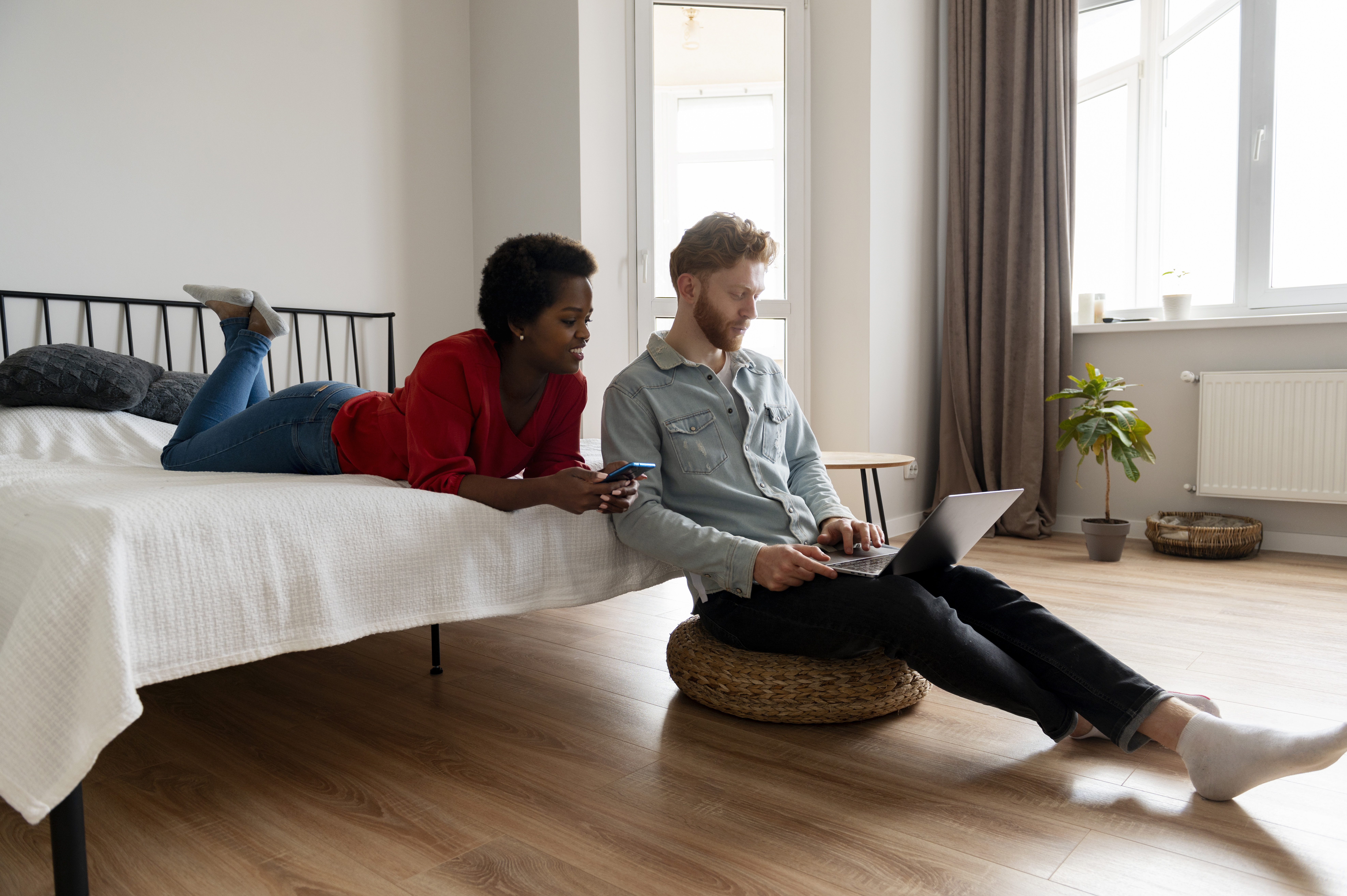 Two people in a bedroom, one lying on a bed with a phone and the other sitting on the floor with a laptop.