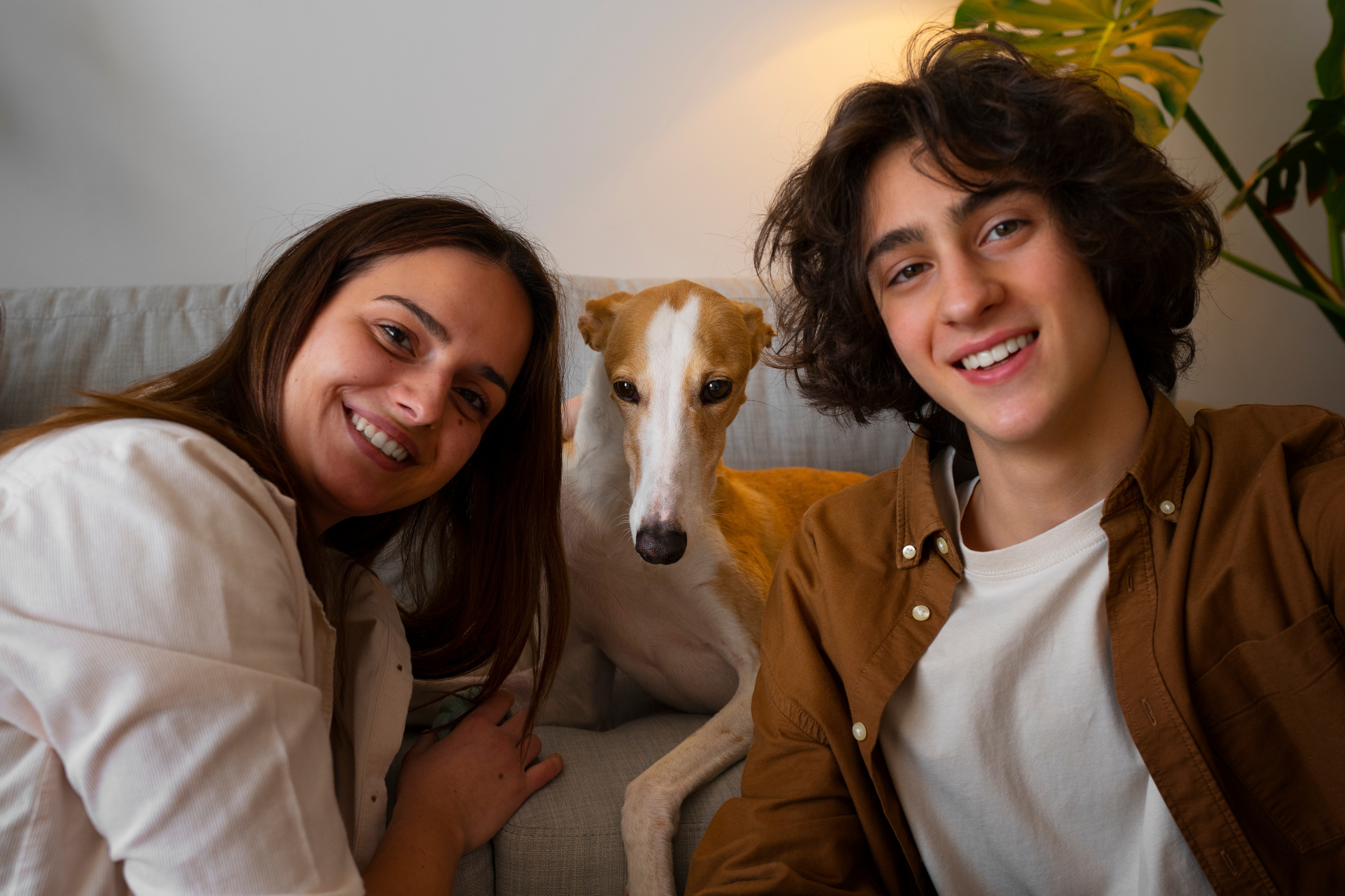 Two smiling people sitting on a couch with a greyhound dog between them.