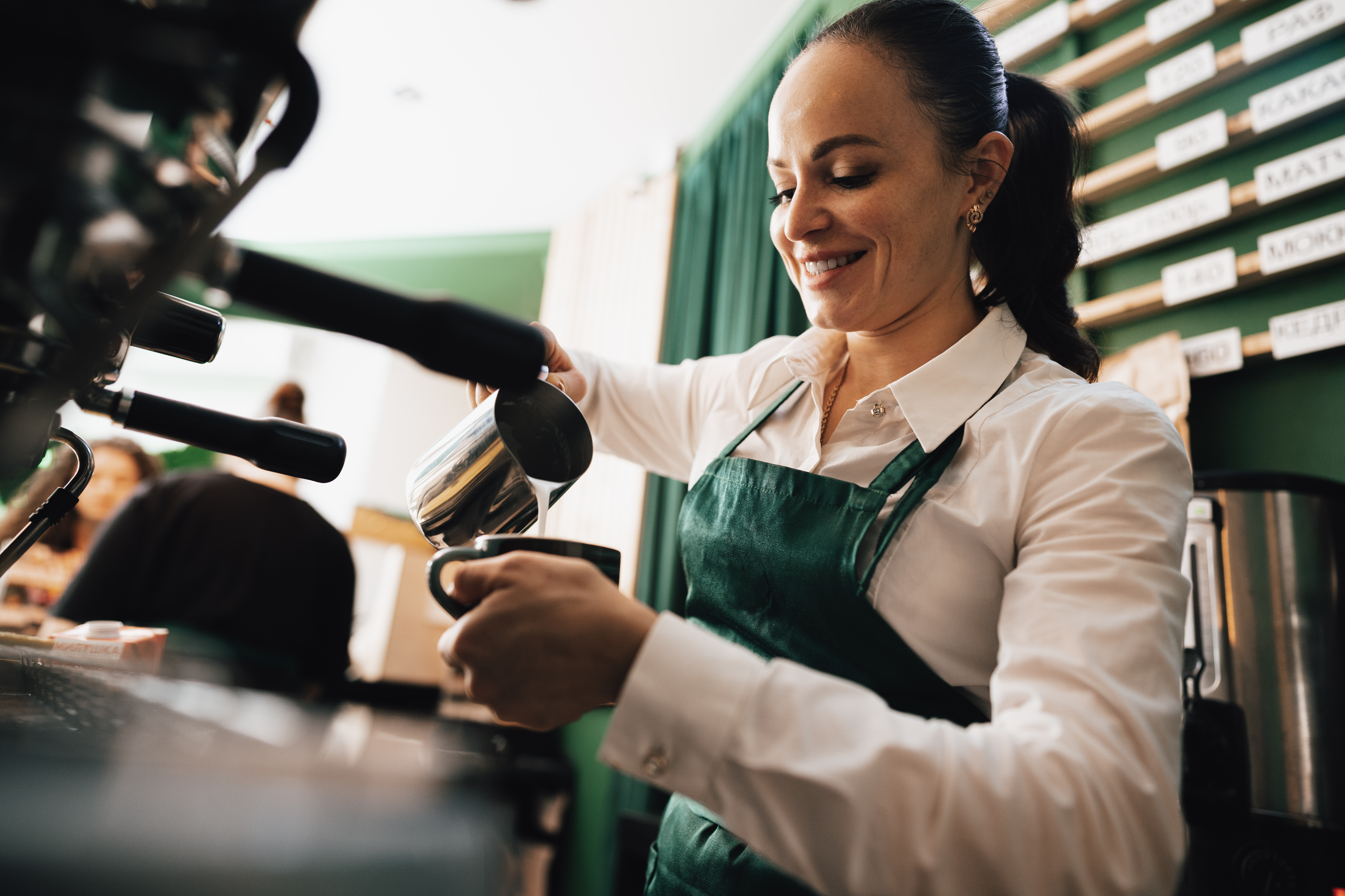 Barista in green apron pouring milk into a coffee cup