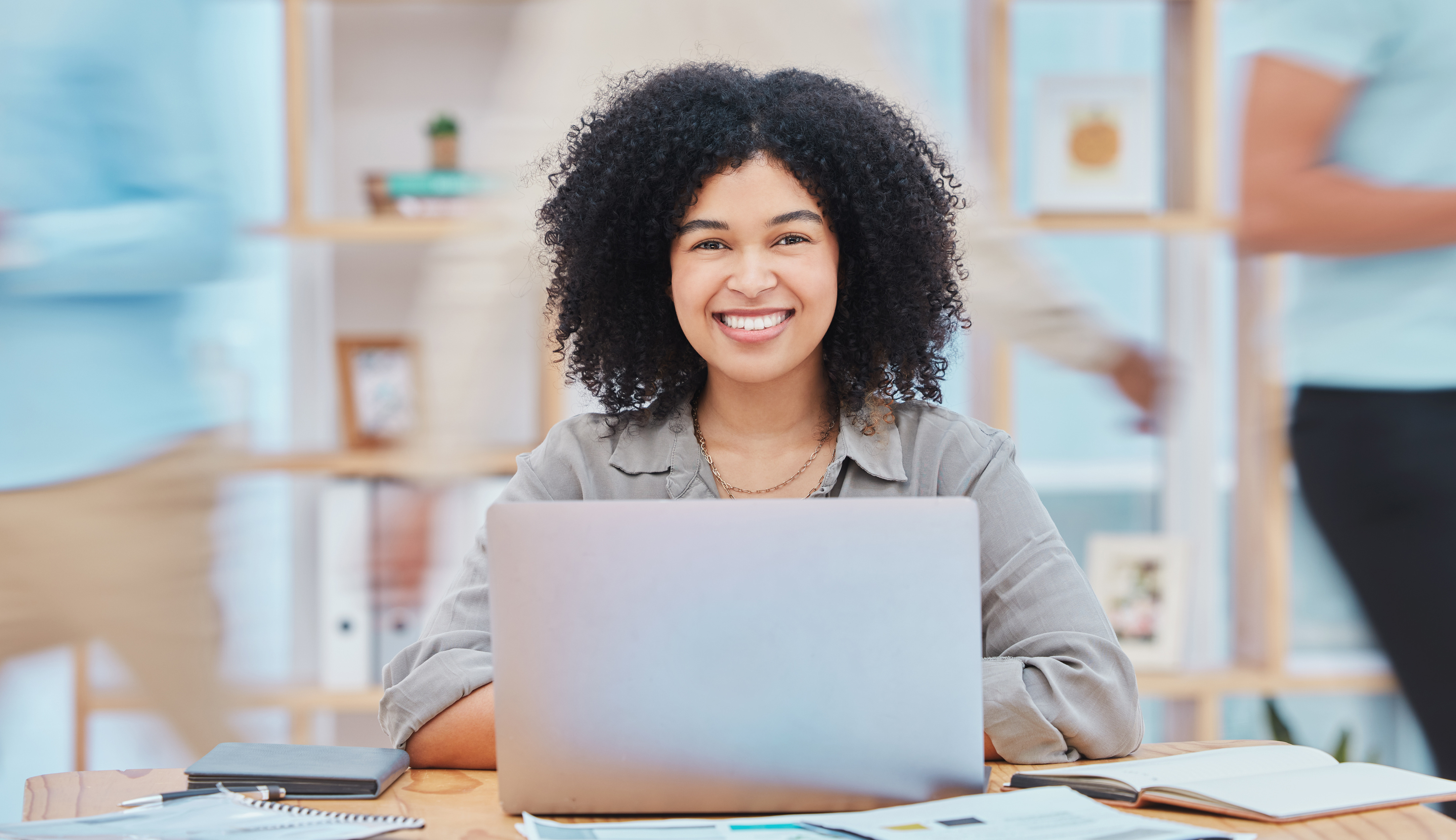 Smiling person sitting at a desk with a laptop in a busy office environment