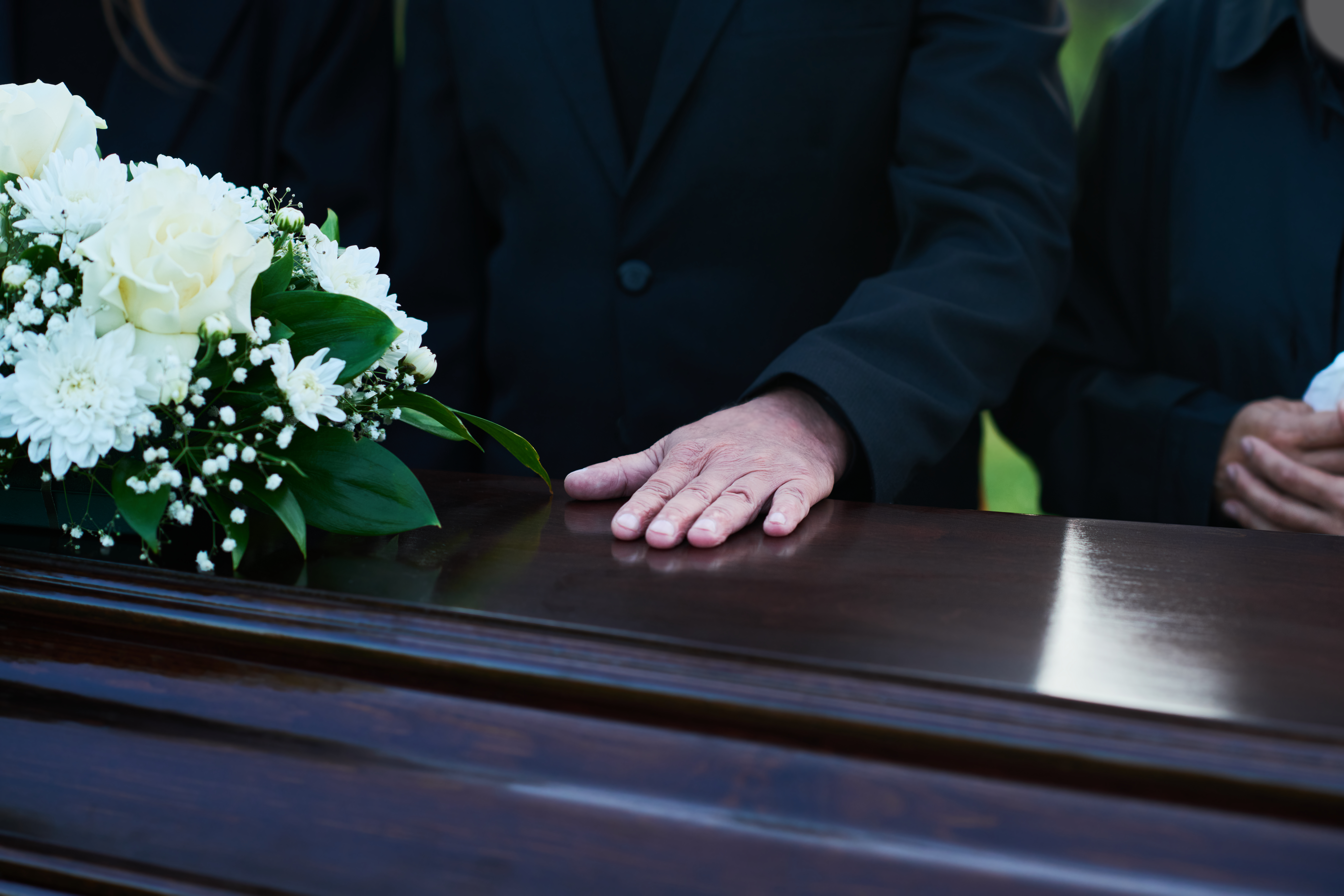 Person's hand resting on a wooden casket with white flowers on top