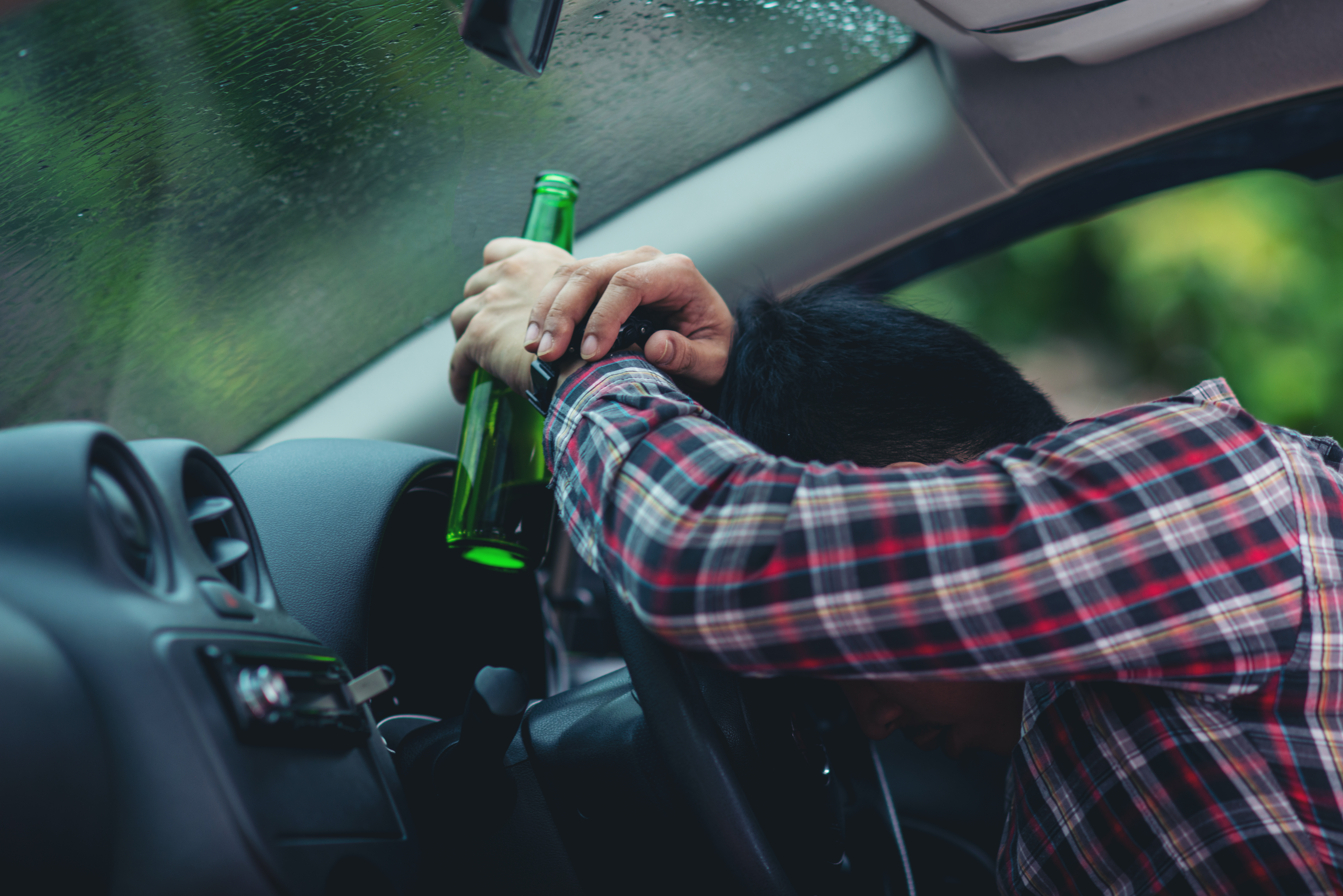 Person in plaid shirt leaning on steering wheel holding a beer bottle