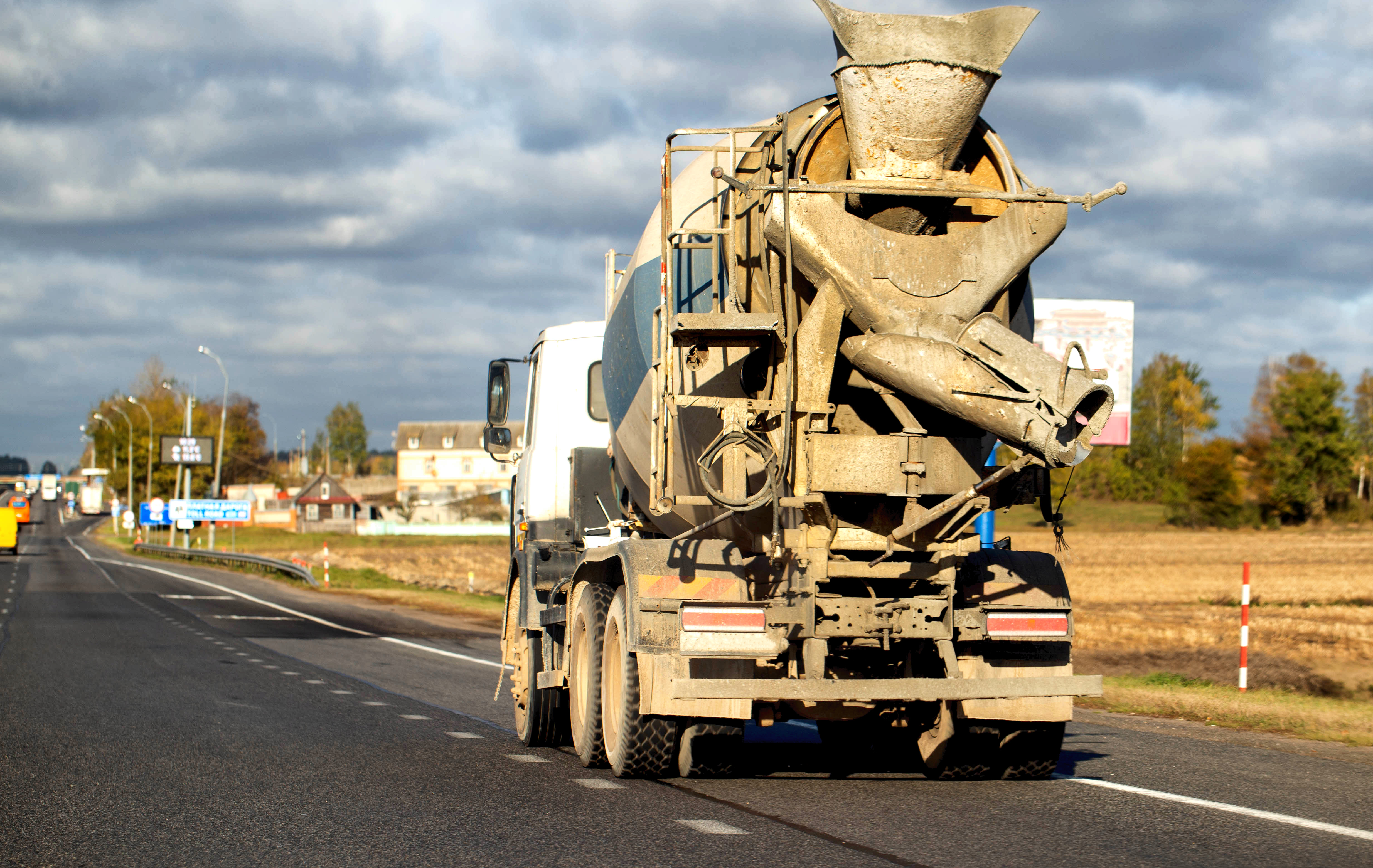 Cement truck driving on a rural road with fields and buildings in the background