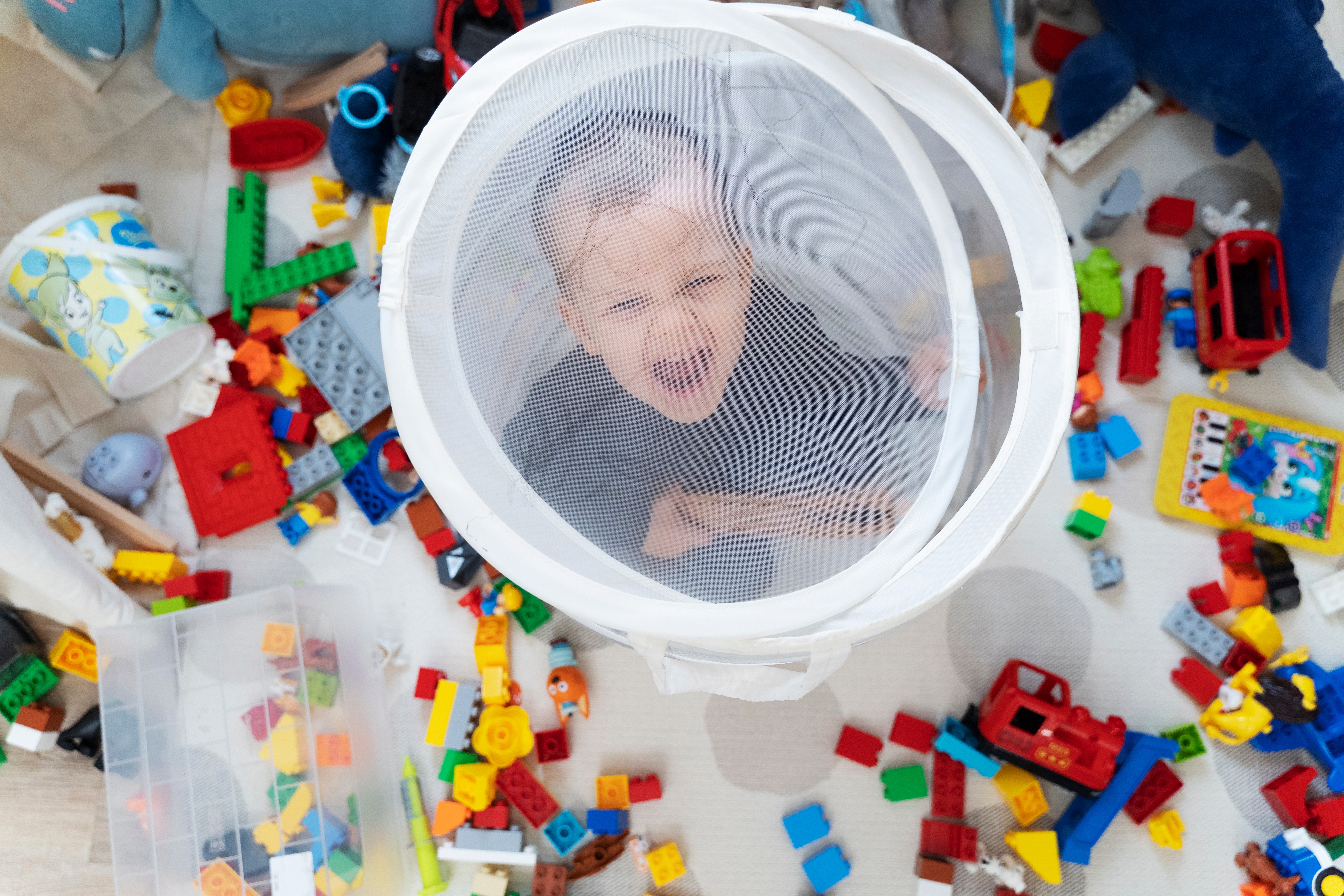 Child playing inside a mesh laundry basket surrounded by colorful building blocks and toys.