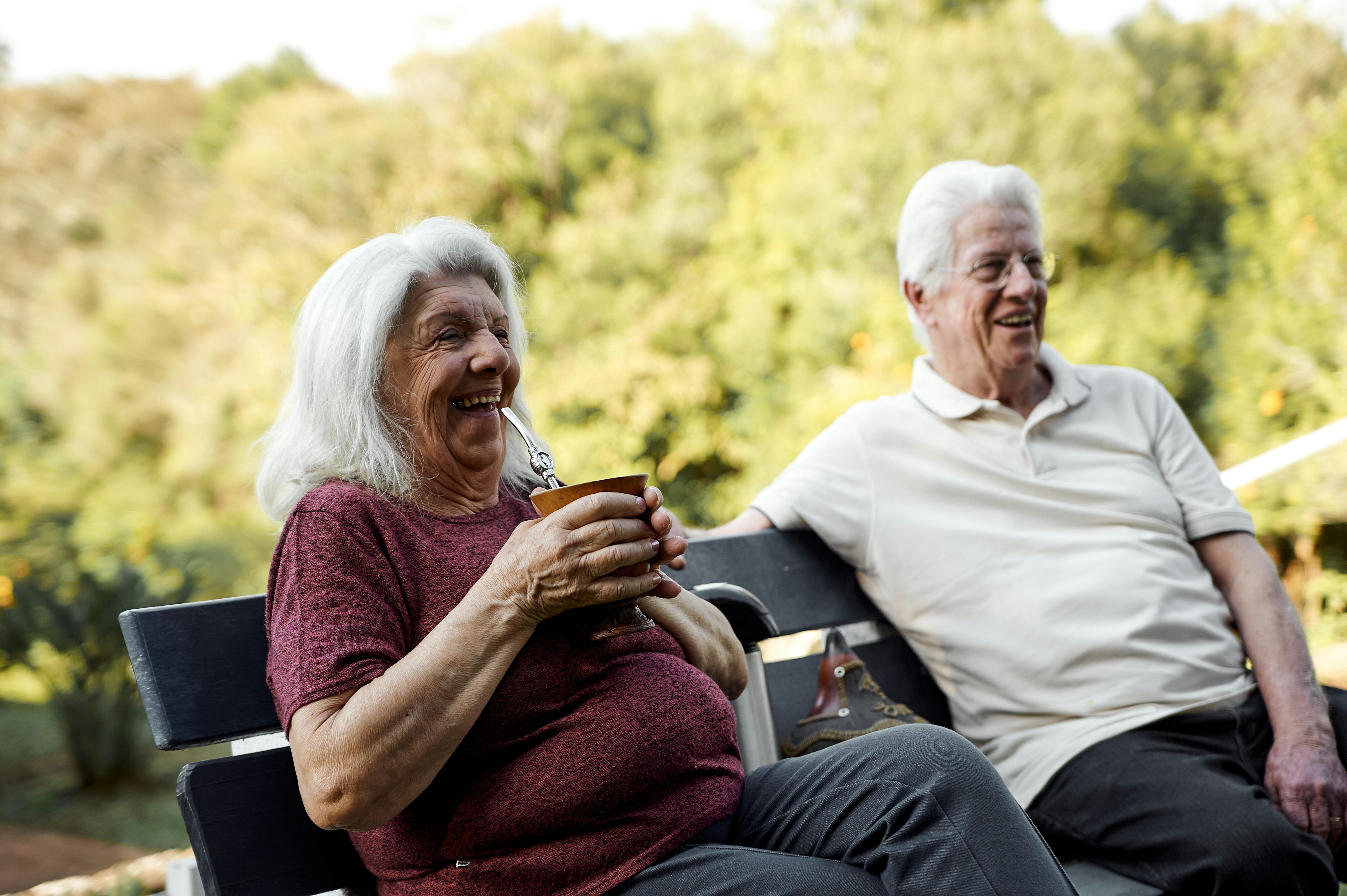 Elderly couple sitting on a bench outdoors, smiling and enjoying a drink.