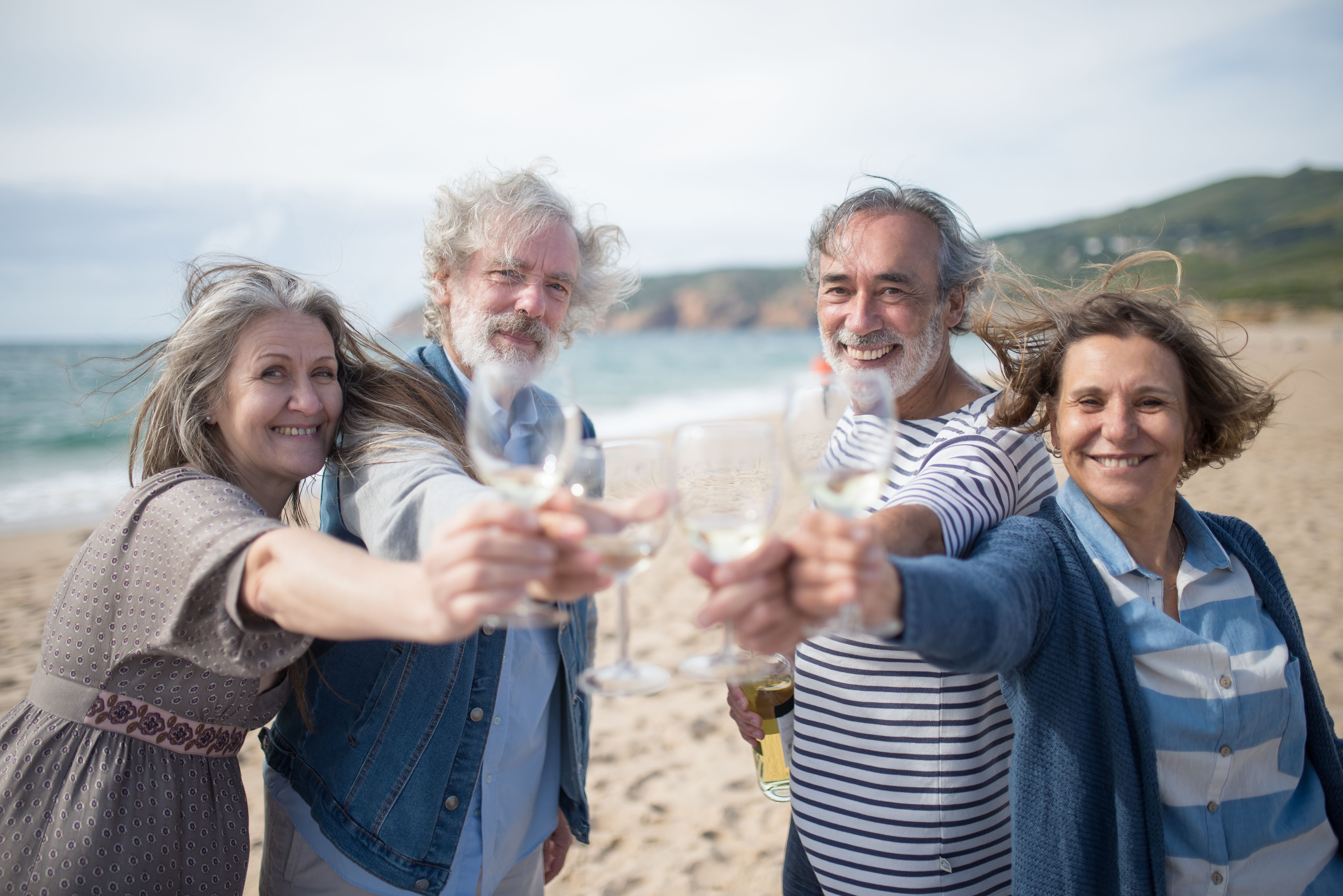 Group of four smiling older adults toasting with wine glasses on a beach.