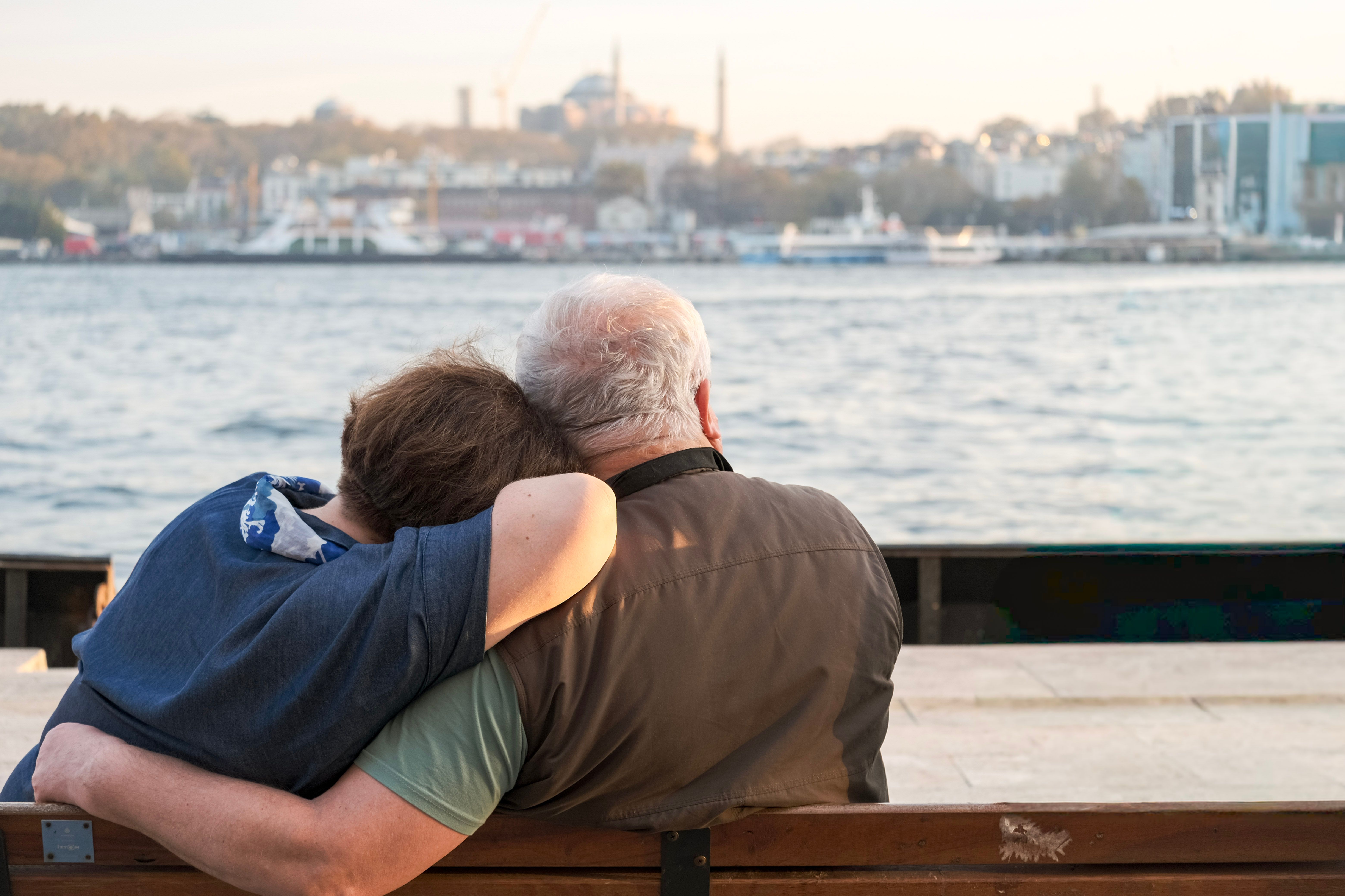 Elderly couple sitting on a bench by the water with a cityscape in the background