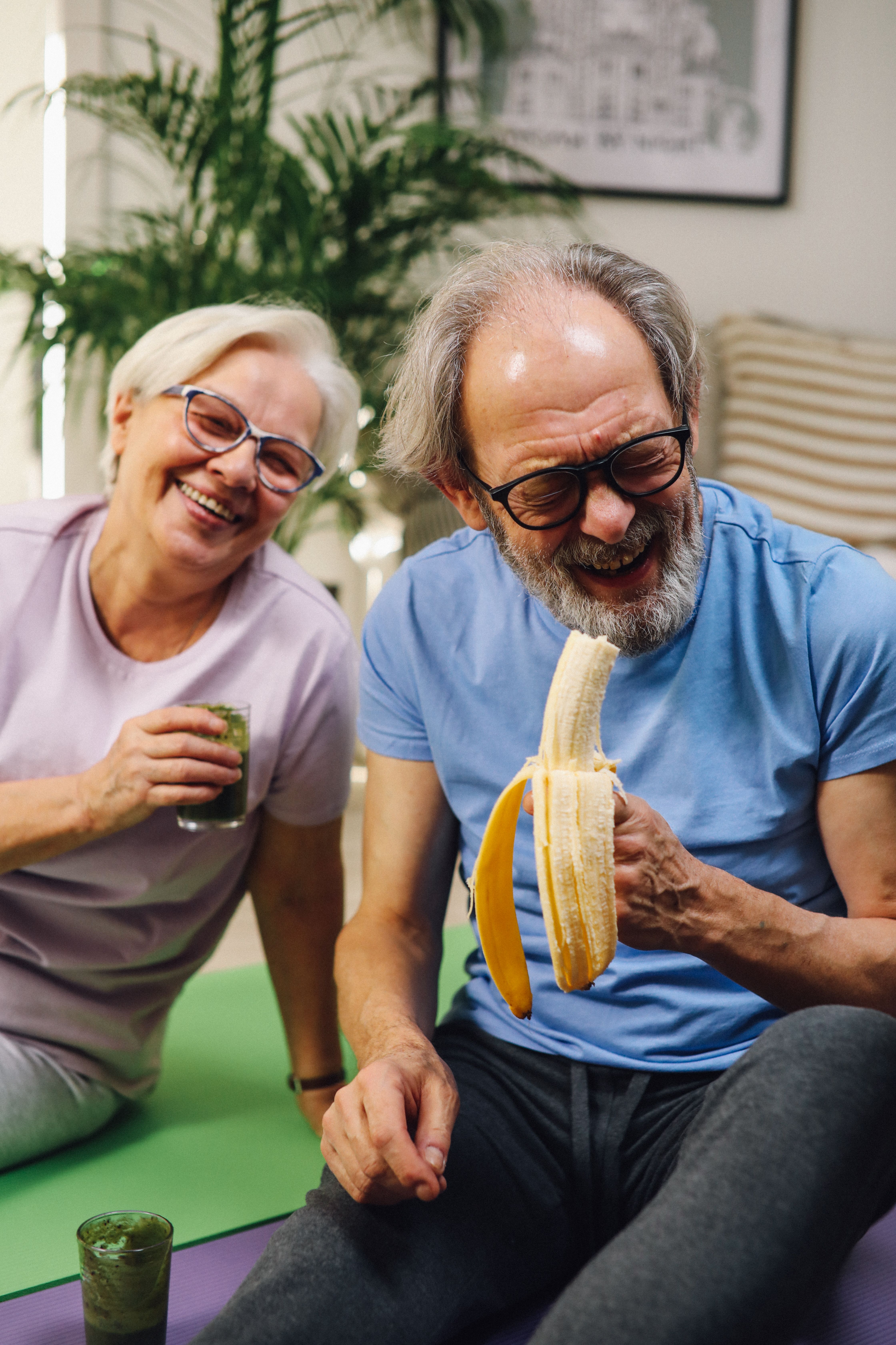 Older couple sitting on yoga mats, smiling and holding a banana and a green smoothie.