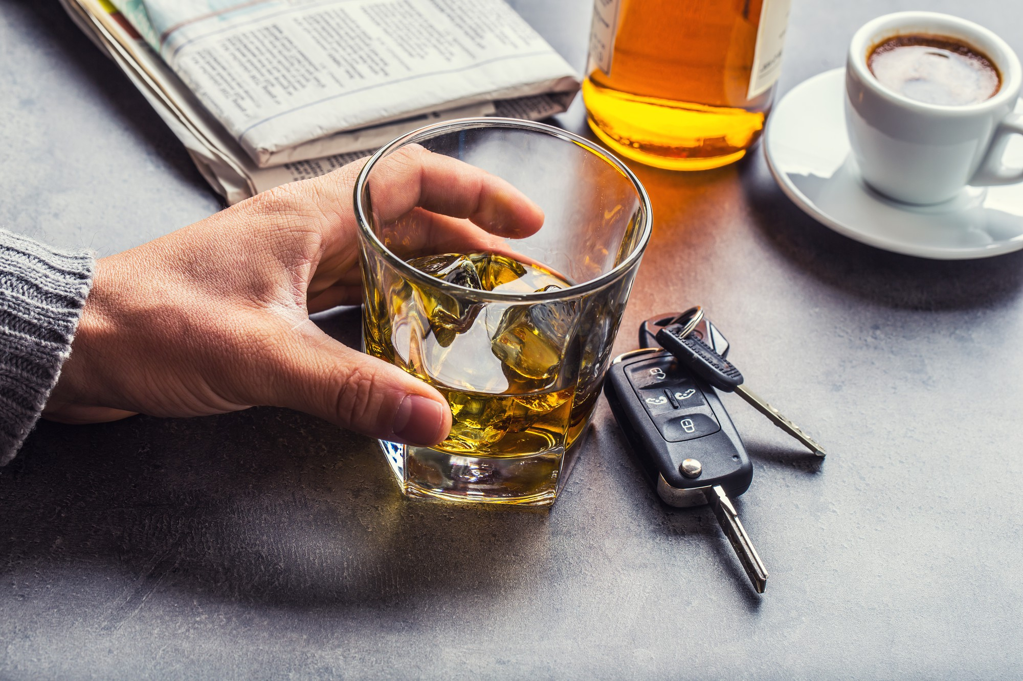 Hand holding a glass of whiskey next to car keys and a coffee cup on a table