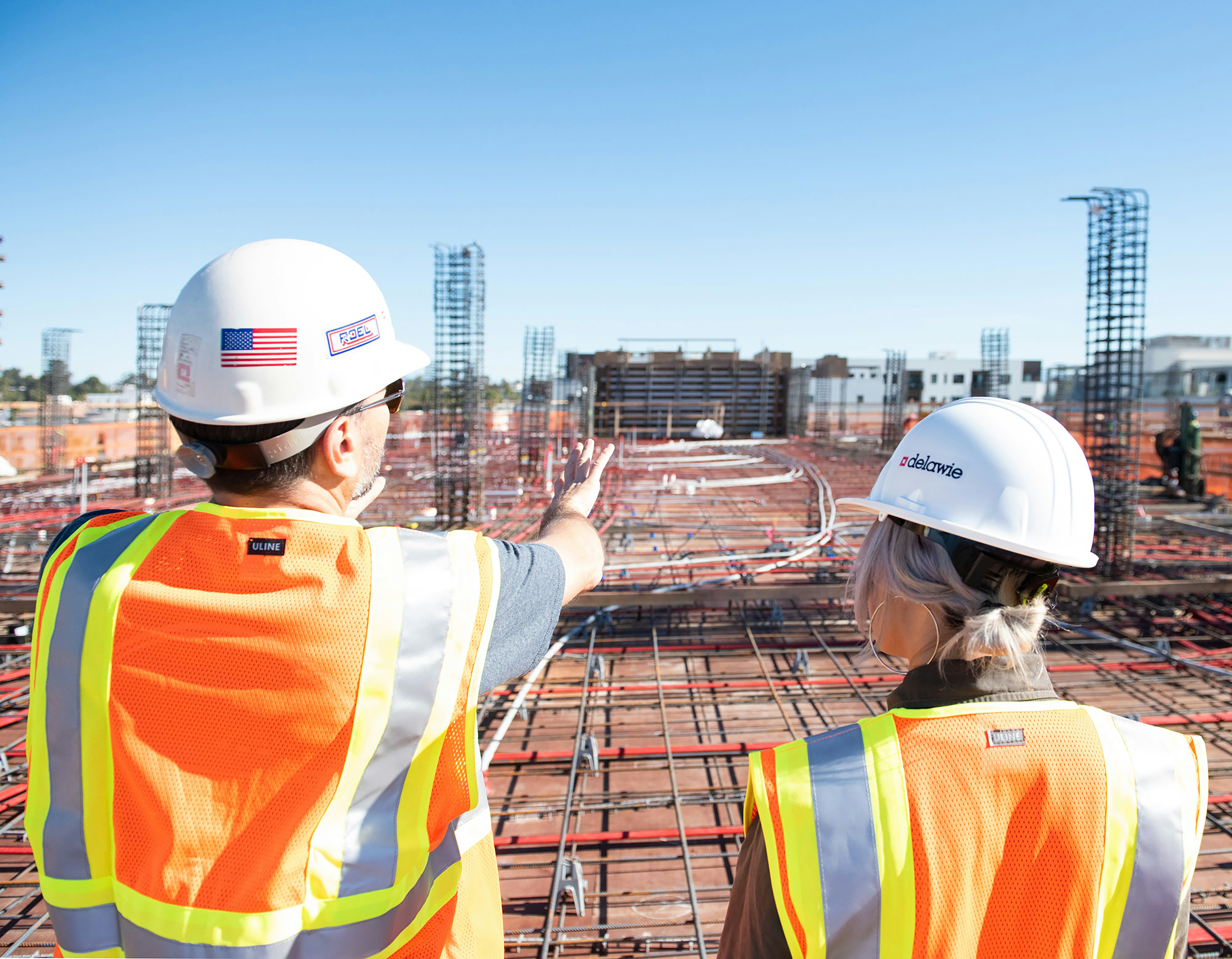 Worker in a yellow safety jacket holds a tablet and looks up in a bright industrial hallway.