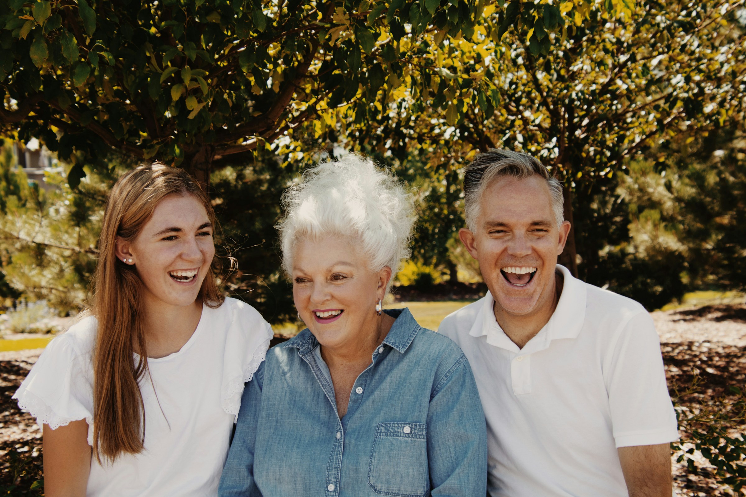 Three people sitting outdoors, smiling and laughing under a tree.