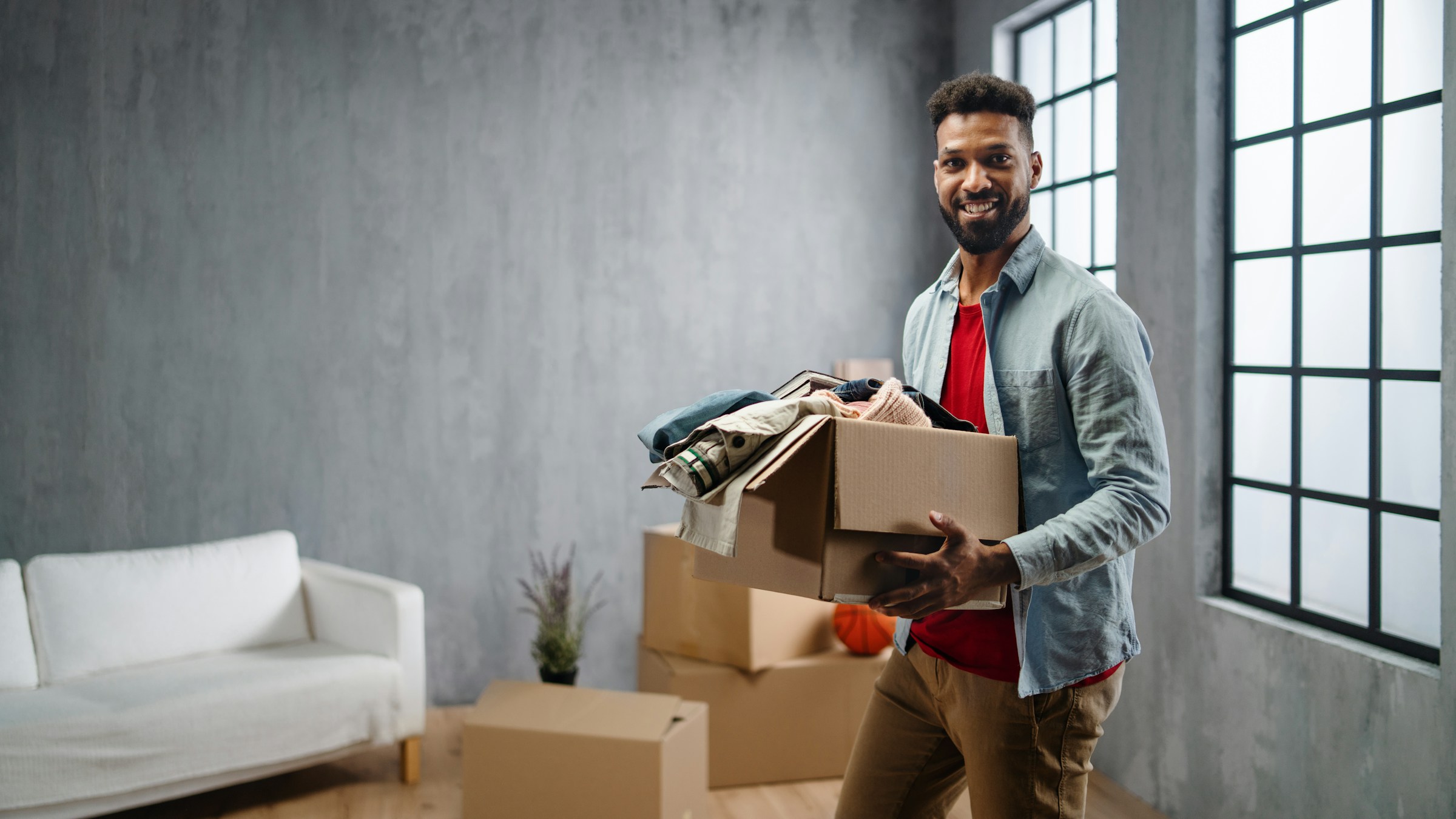 Man smiling while carrying a cardboard box in a room with moving boxes.