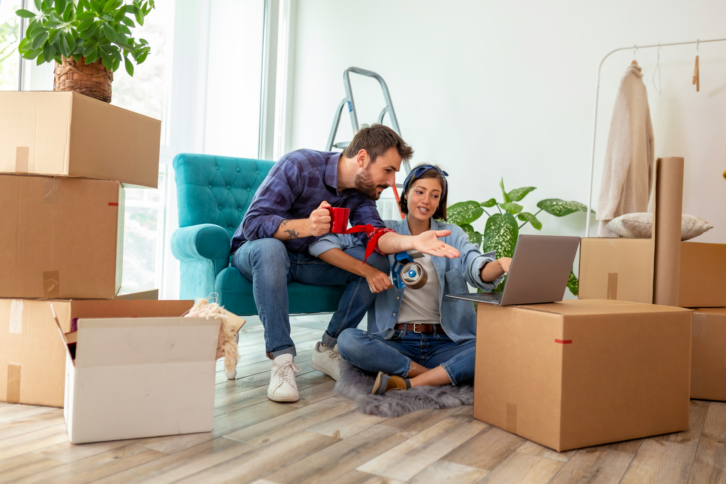 Couple sitting among moving boxes, looking at a laptop and planning.
