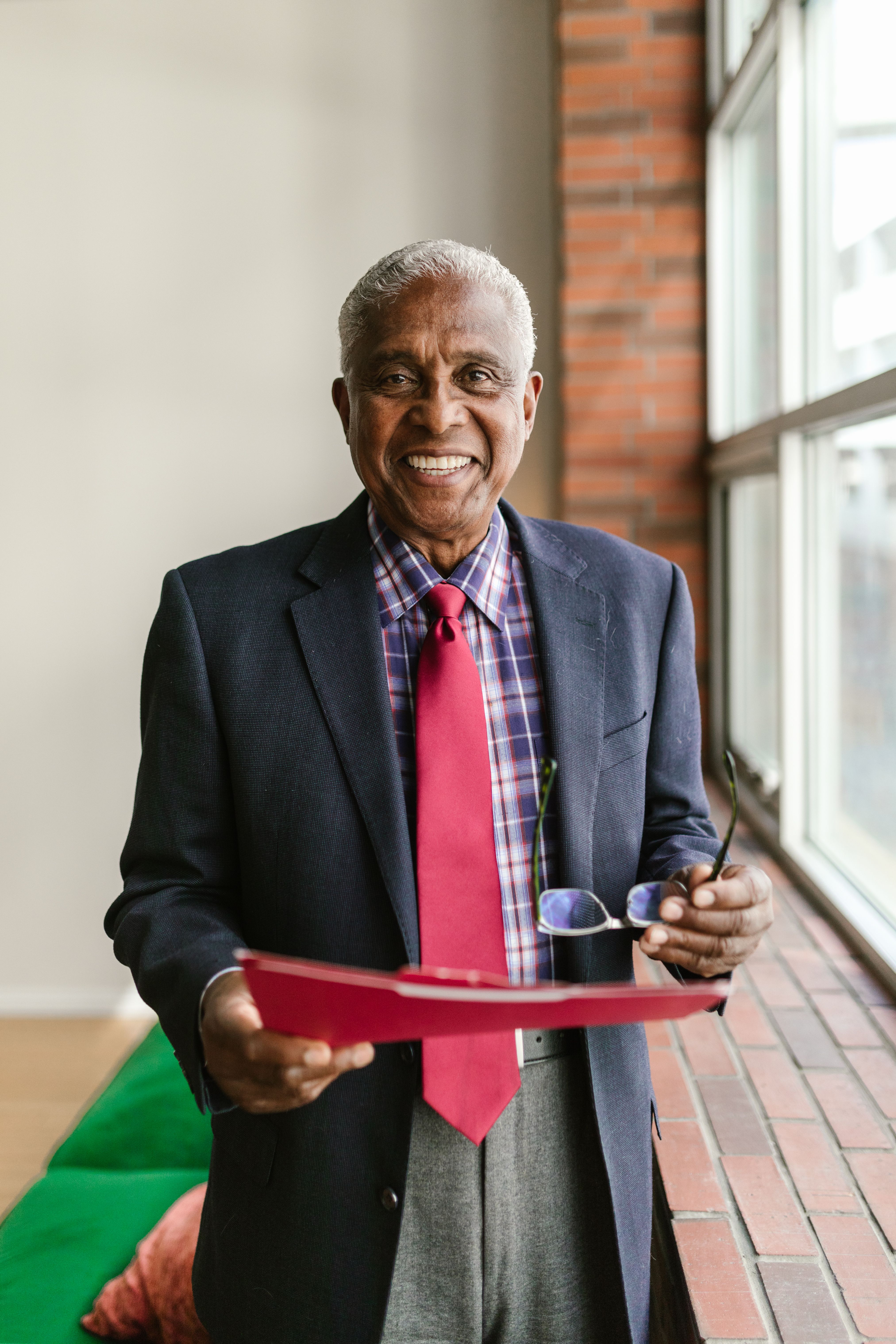Elderly man in a suit holding a folder and glasses, standing by a window.