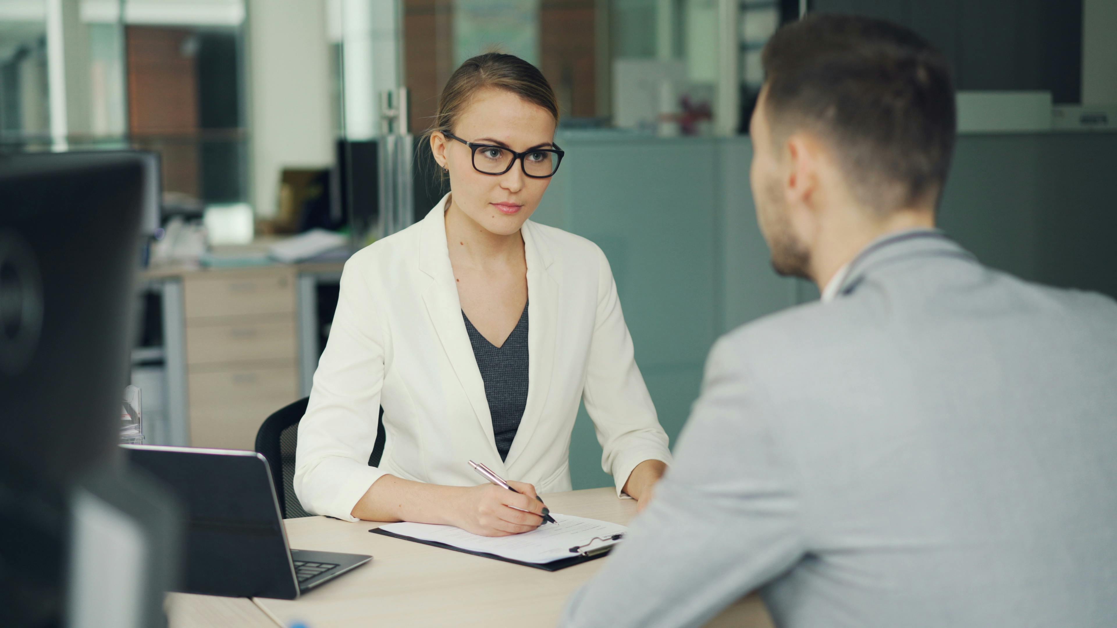 Business meeting between a woman in a white blazer and a man in a gray suit at an office desk.