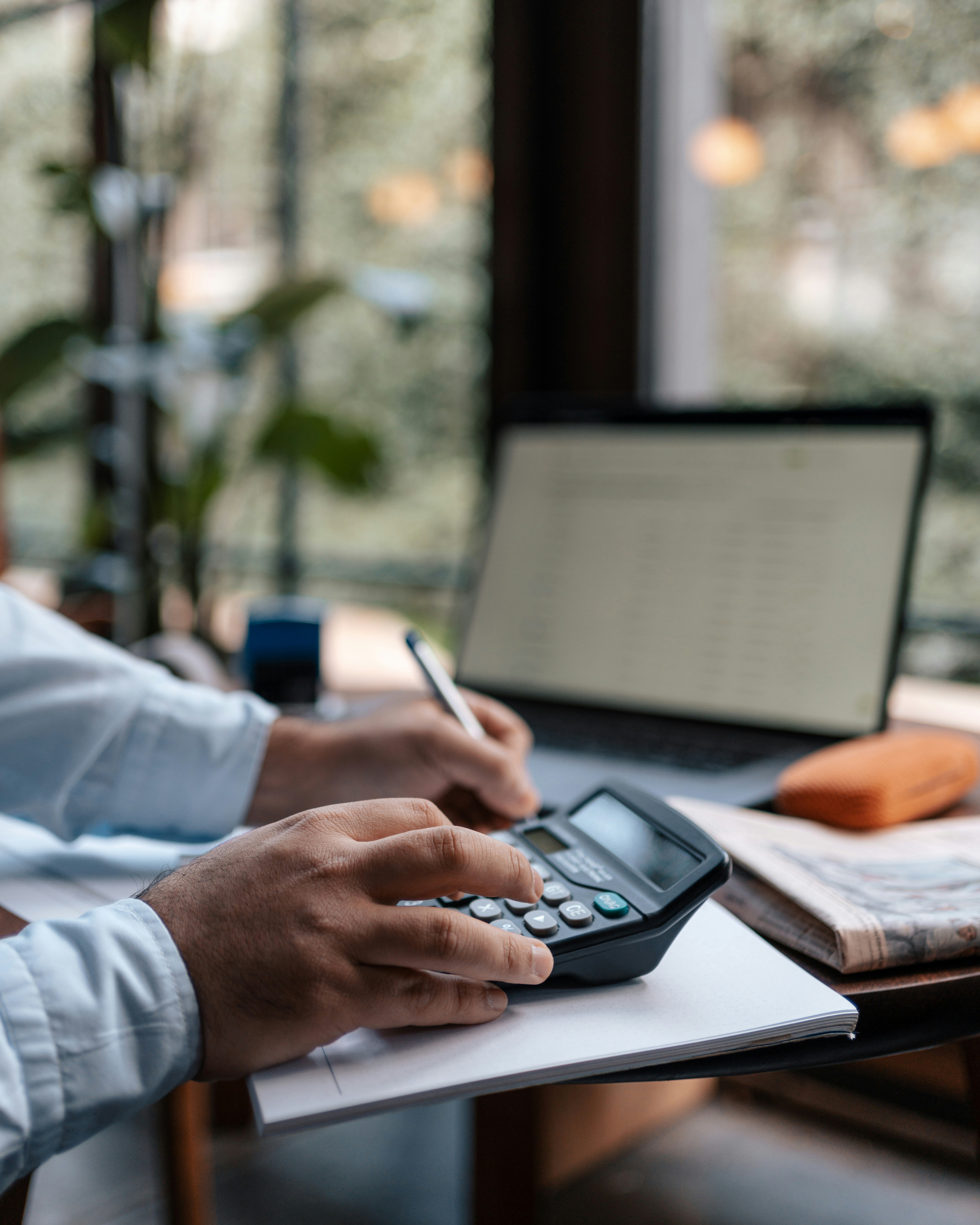 Person using a calculator and writing on a notepad at a desk with a laptop.