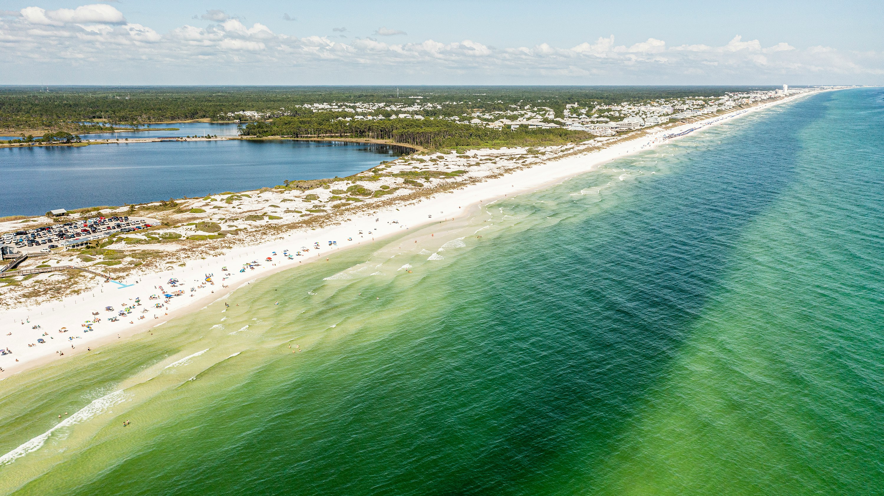 Aerial view of a long sandy beach with clear green water and scattered beachgoers.