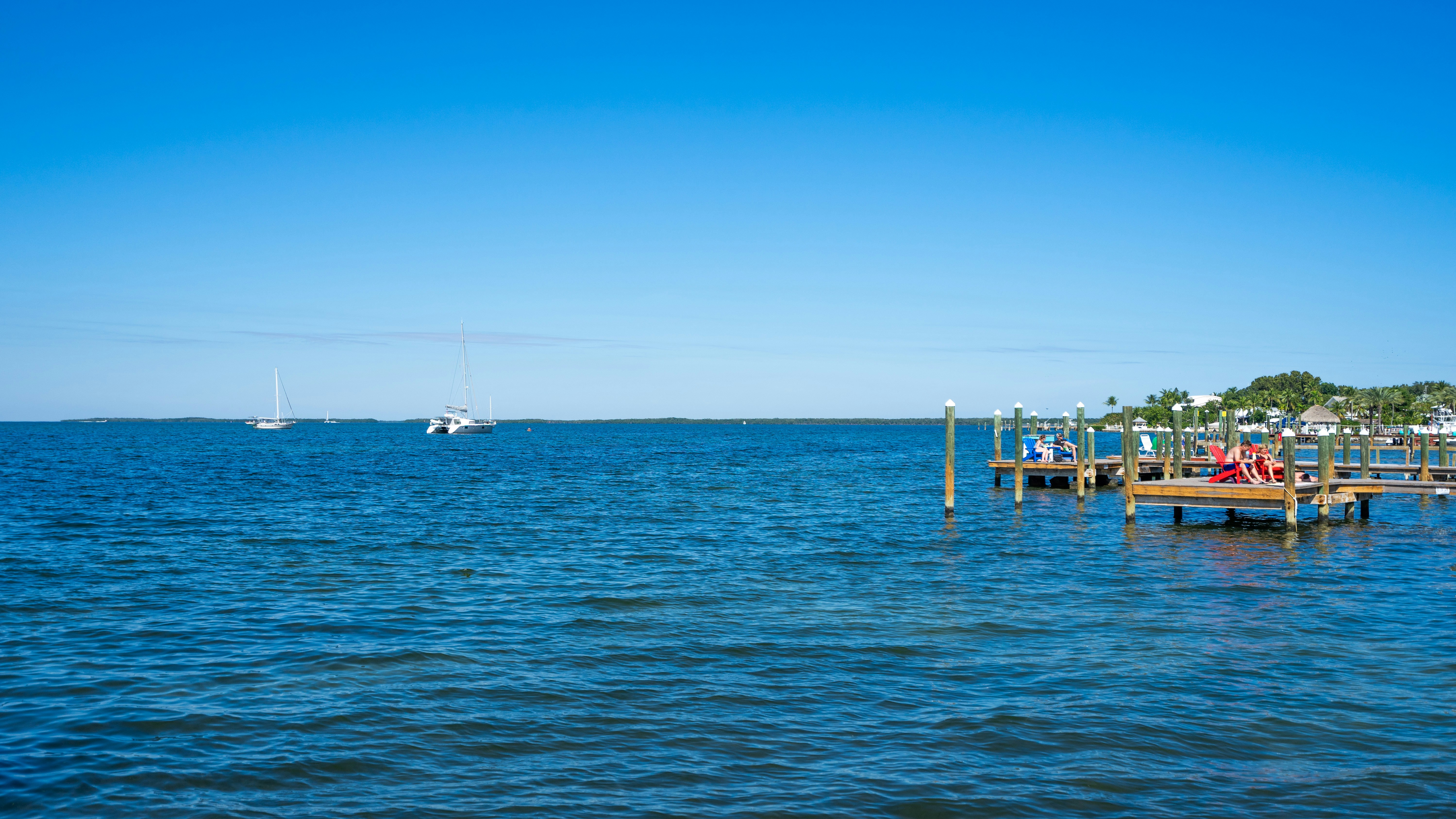 Scenic view of a calm blue sea with sailboats and a wooden dock.