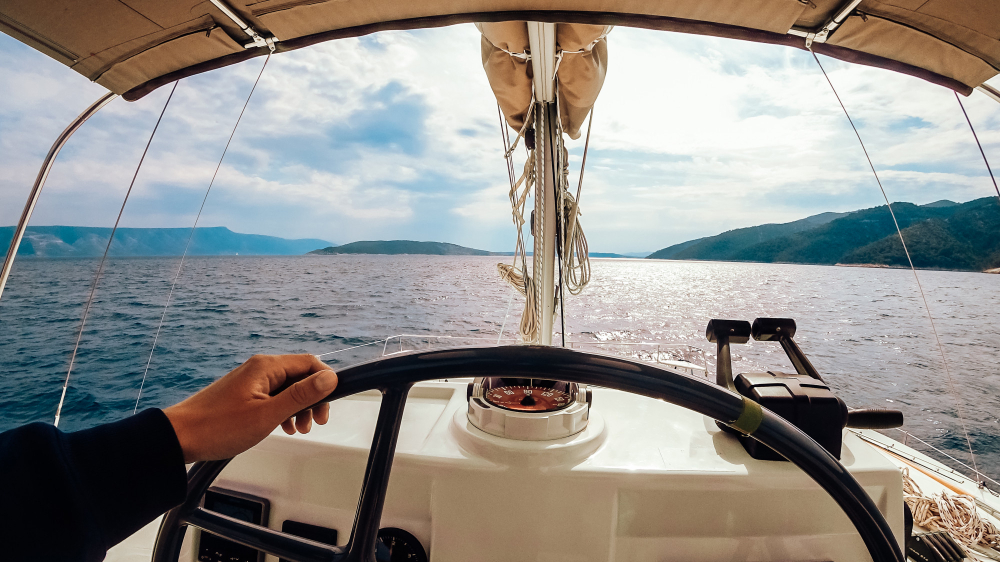 Person steering a sailboat on a sunny day with mountains in the background