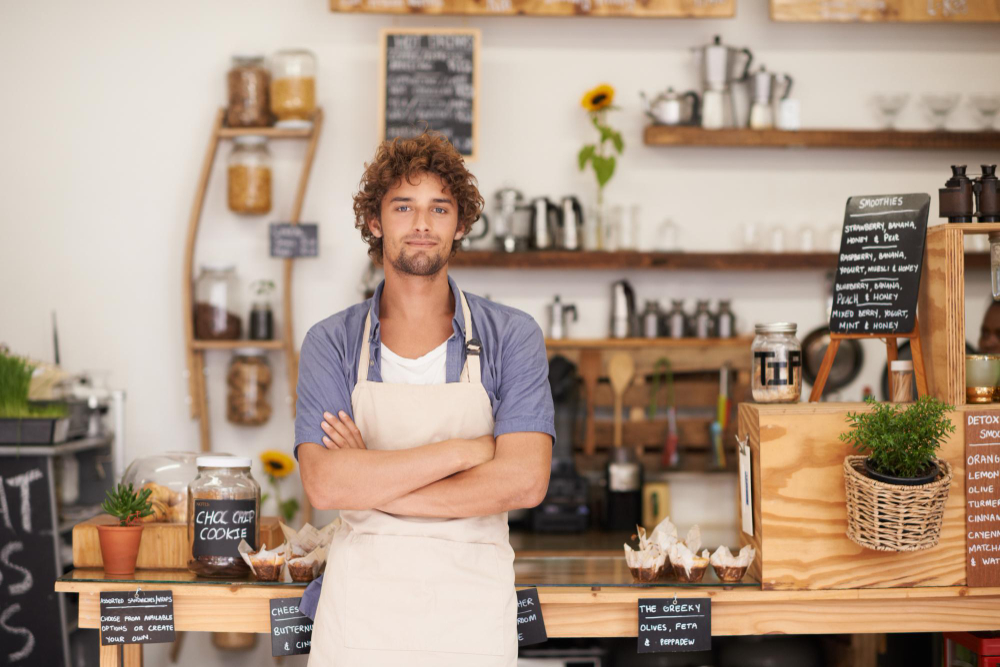 Man with curly hair wearing an apron standing in a cafe with arms crossed