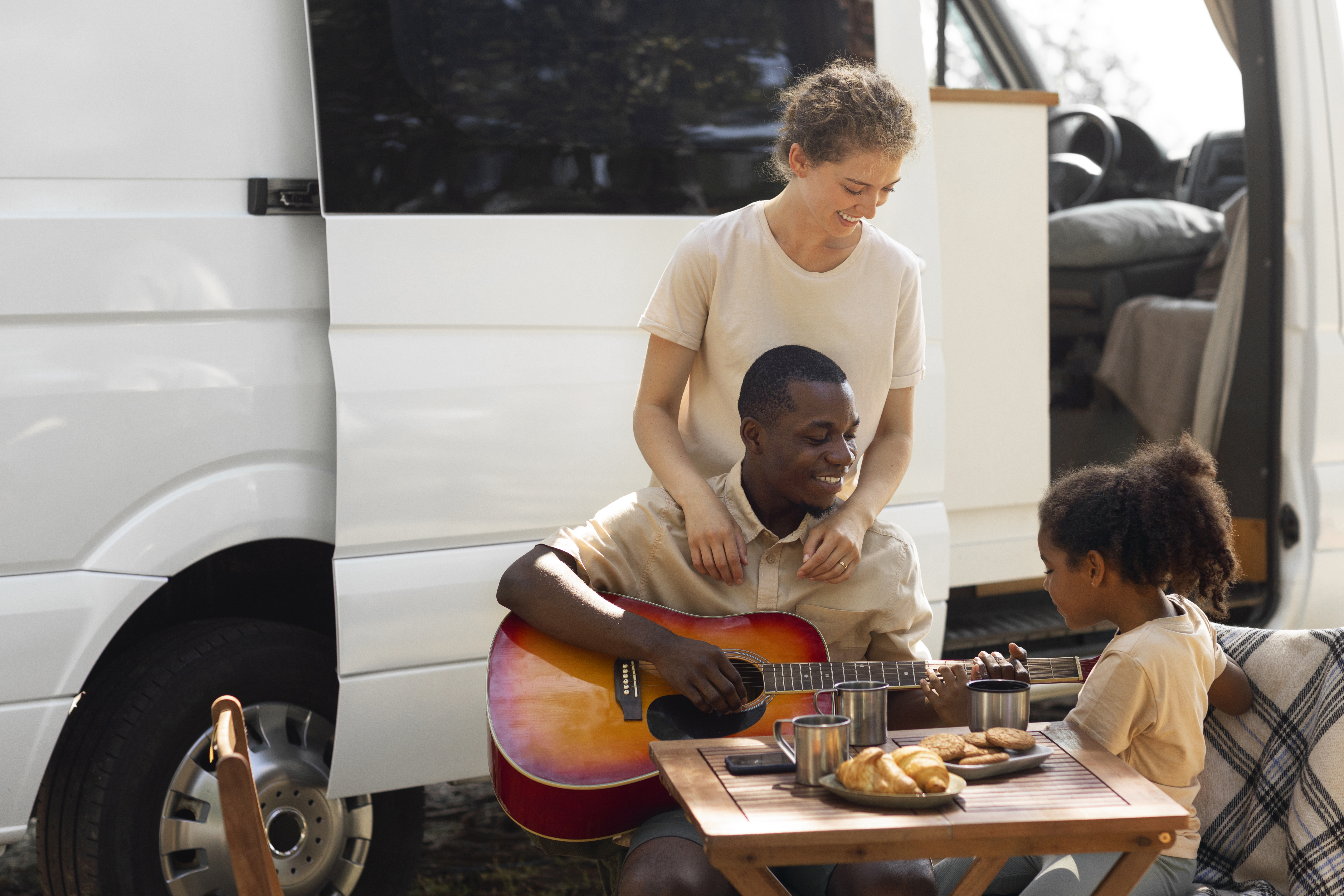 Family enjoying a picnic by a camper van with a man playing guitar.