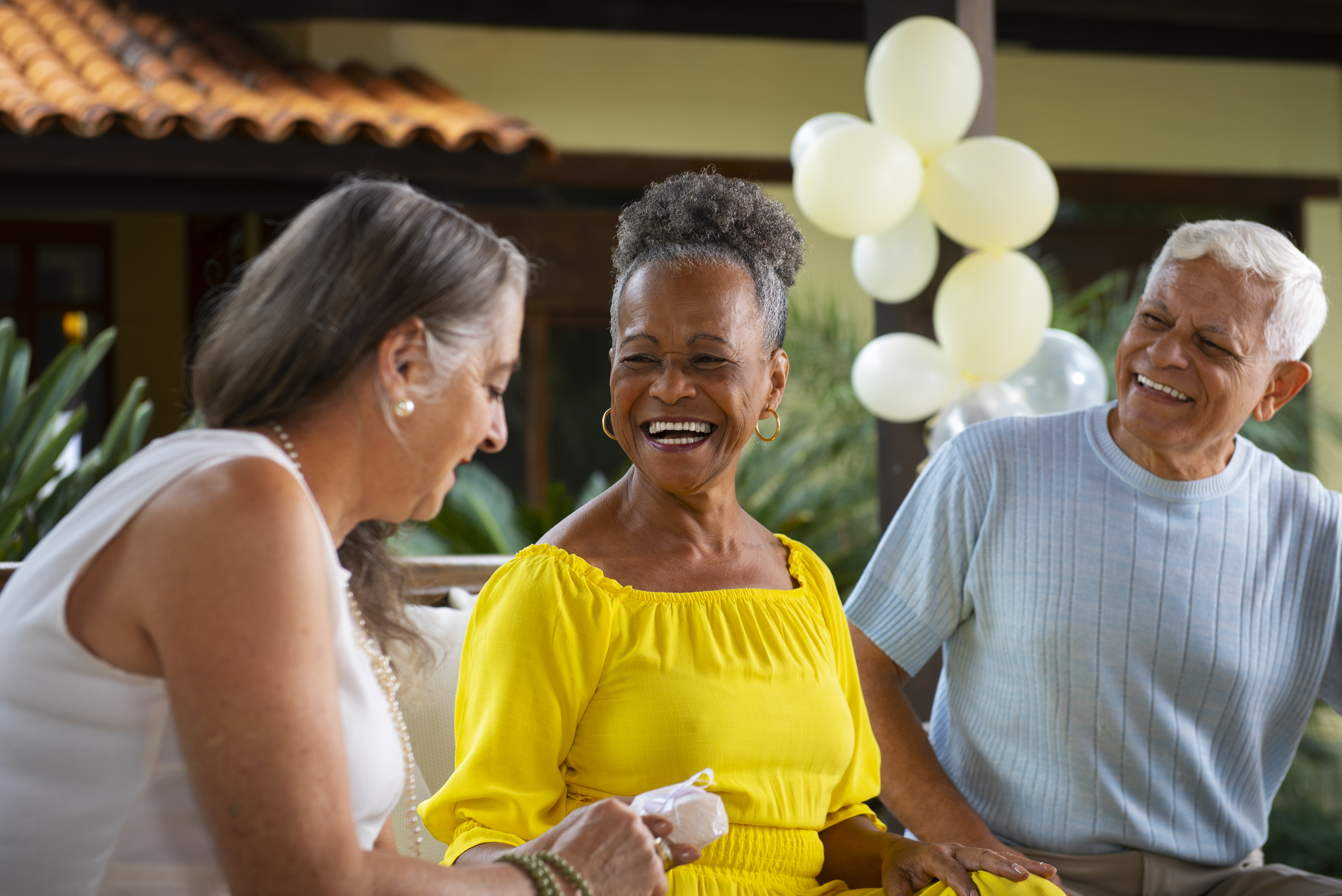 Three older adults smiling and talking at an outdoor gathering with balloons in the background.