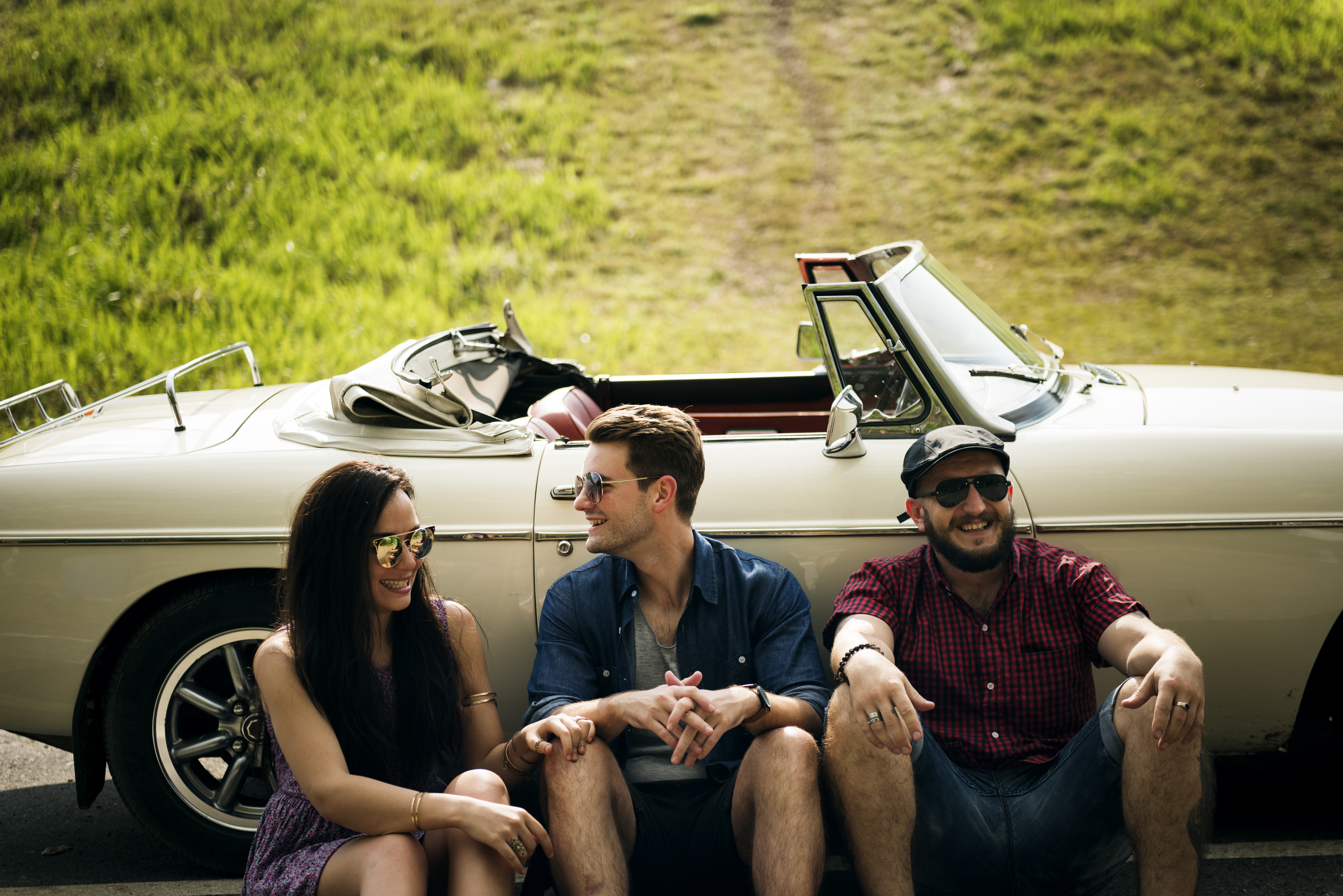 Three people sitting in front of a vintage convertible car on a sunny day.