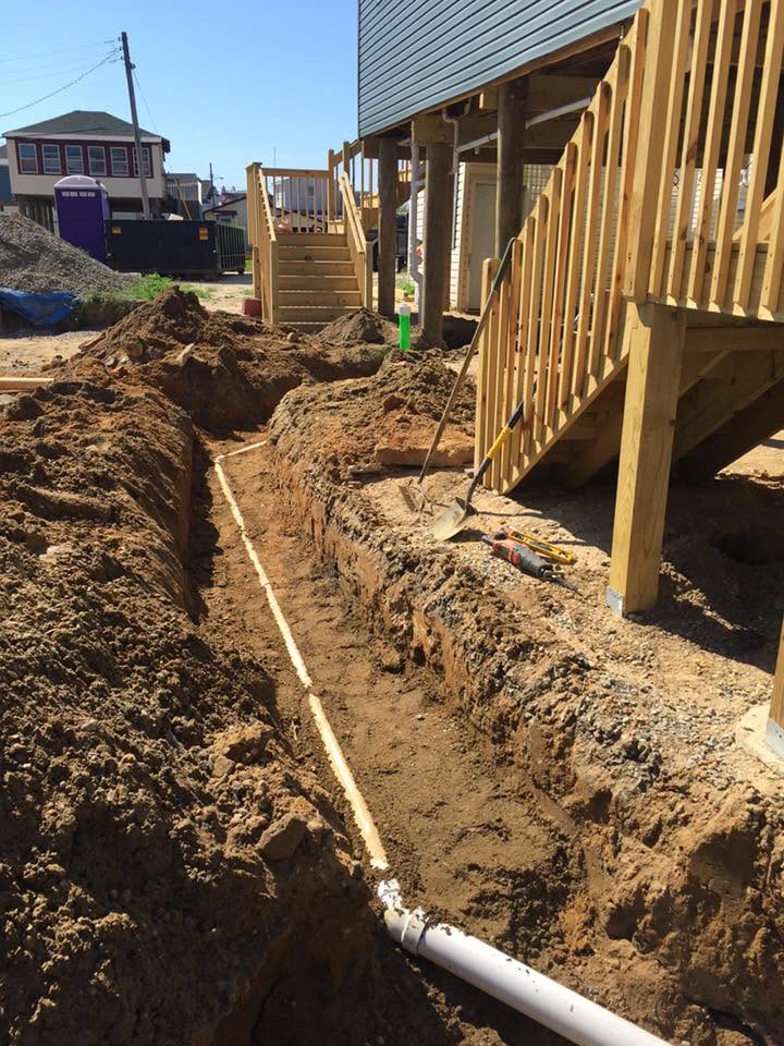 Trench with exposed plumbing pipes next to a house under construction